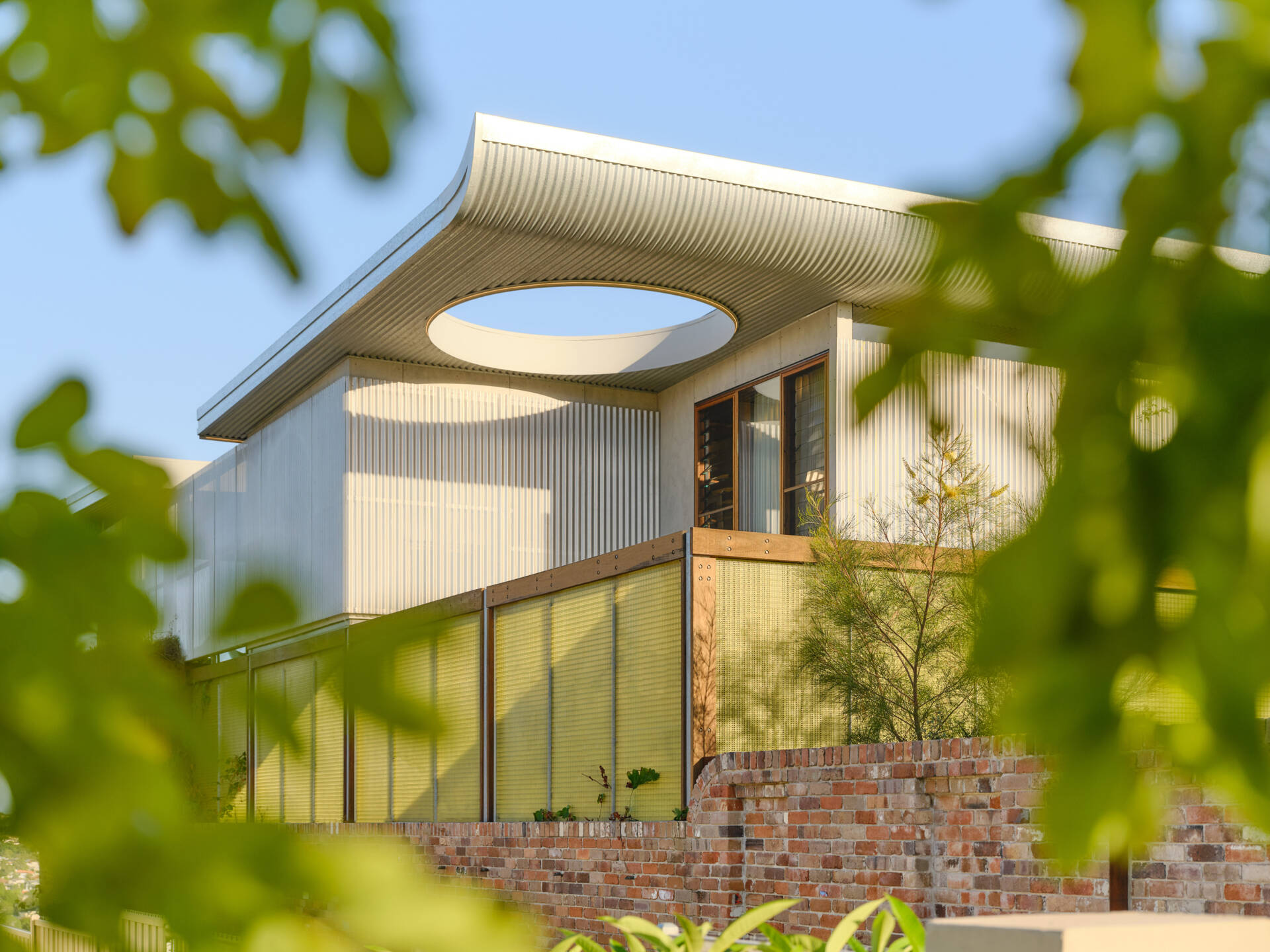 A stunning Sydney home featuring a dramatic butterfly roof, circular windows, curved brickwork, and translucent jade-green screens that create privacy without blocking light.