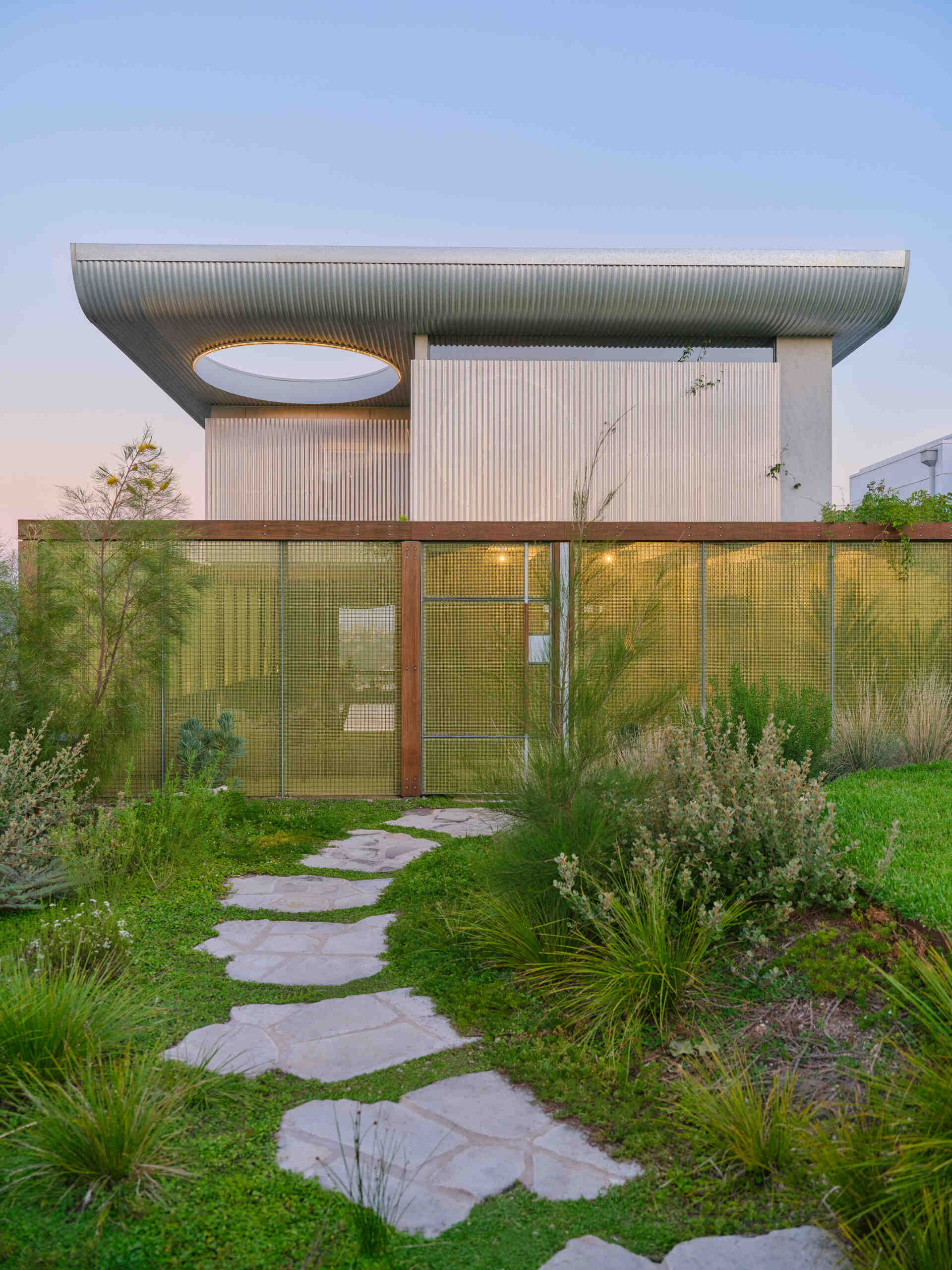 This garden entry path winds through lush planting and translucent screens, creating a welcoming arrival experience filled with greenery and filtered light.