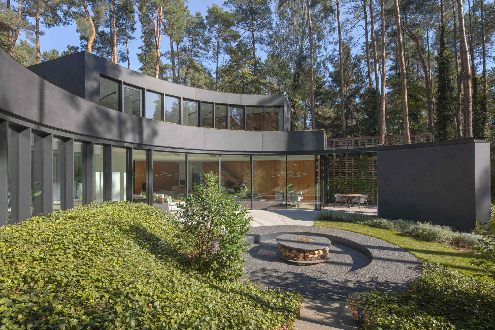 Curved glass walls wrap around a private courtyard in House with a Glade. Custom arched glazing, sliding glass doors, and a central fire pit create a dramatic connection between architecture, light, and the surrounding forest.