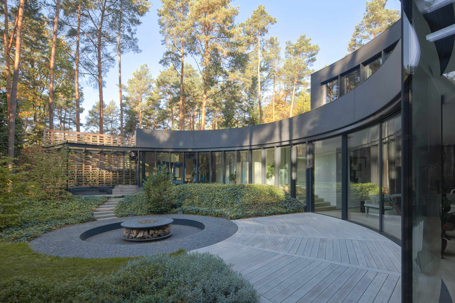 Curved glass walls wrap around a private courtyard in House with a Glade. Custom arched glazing, sliding glass doors, and a central fire pit create a dramatic connection between architecture, light, and the surrounding forest.