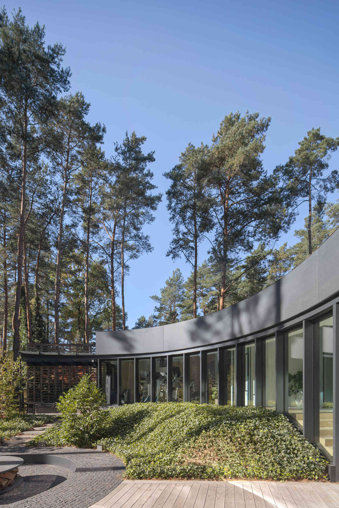Curved glass walls wrap around a private courtyard in House with a Glade. Custom arched glazing, sliding glass doors, and a central fire pit create a dramatic connection between architecture, light, and the surrounding forest.