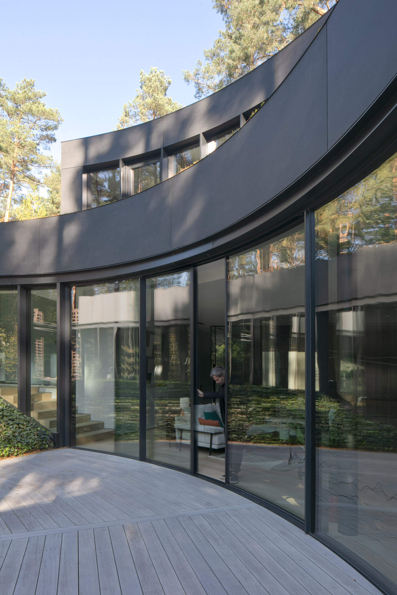 Curved glass walls wrap around a private courtyard in House with a Glade. Custom arched glazing, sliding glass doors, and a central fire pit create a dramatic connection between architecture, light, and the surrounding forest.