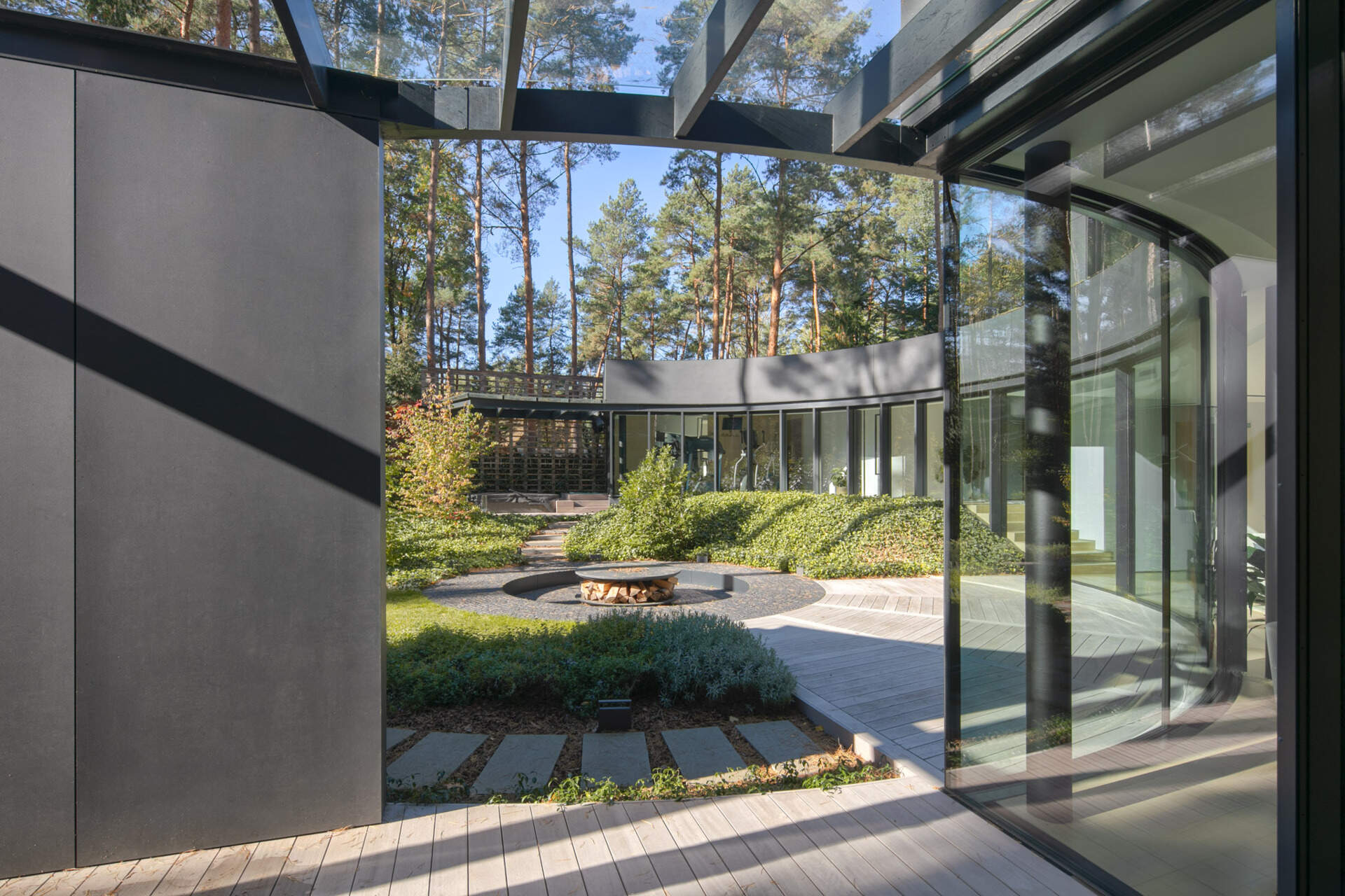 Curved glass walls wrap around a private courtyard in House with a Glade. Custom arched glazing, sliding glass doors, and a central fire pit create a dramatic connection between architecture, light, and the surrounding forest.