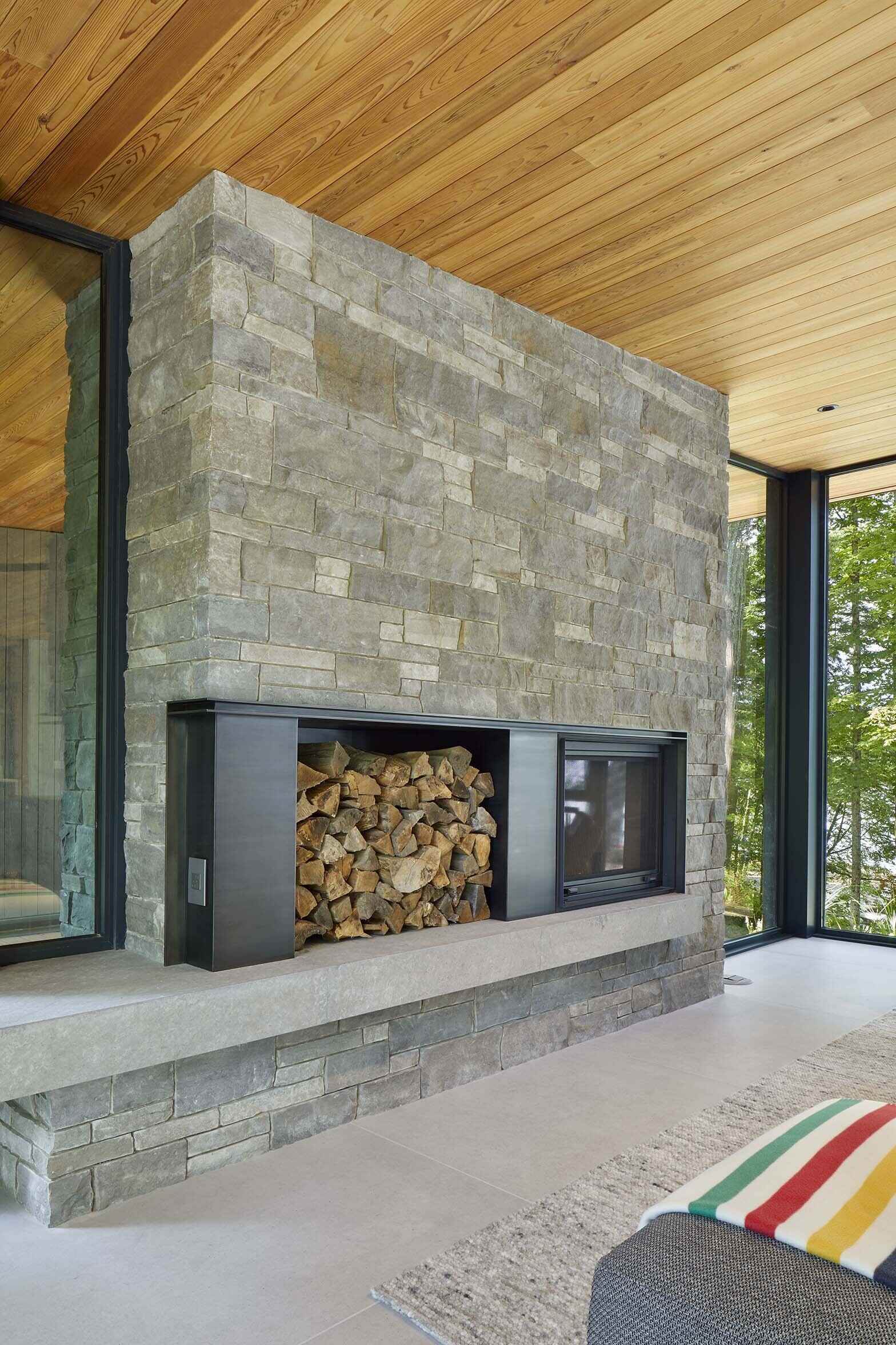 The central great room contains the living space and is defined by expansive floor-to-ceiling glazing that frames views across Percy Lake. A blackened-steel wood-burning fireplace with a stone surround, anchors the room and acts as a focal point within the open space.