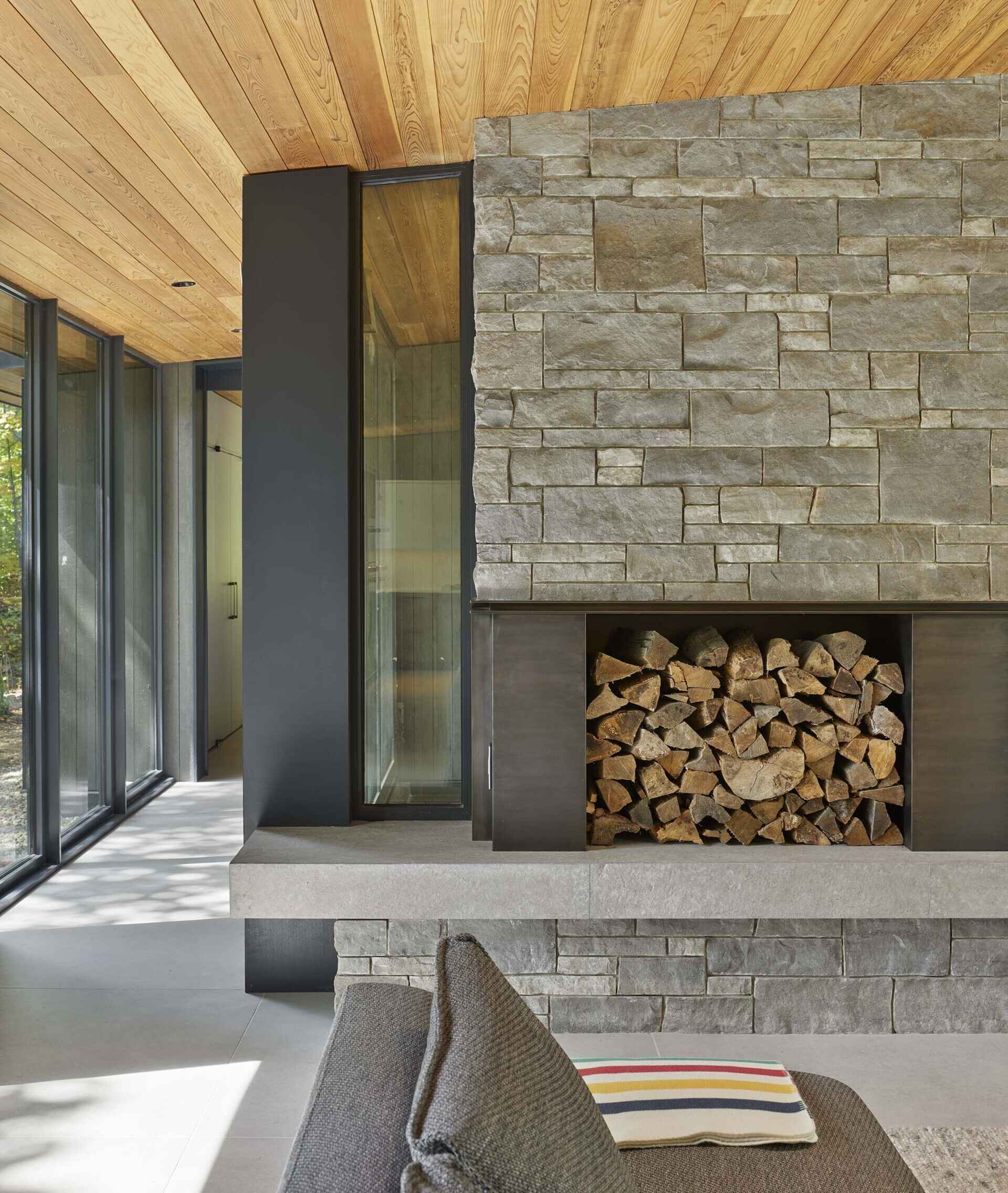 The central great room contains the living space and is defined by expansive floor-to-ceiling glazing that frames views across Percy Lake. A blackened-steel wood-burning fireplace with a stone surround, anchors the room and acts as a focal point within the open space.