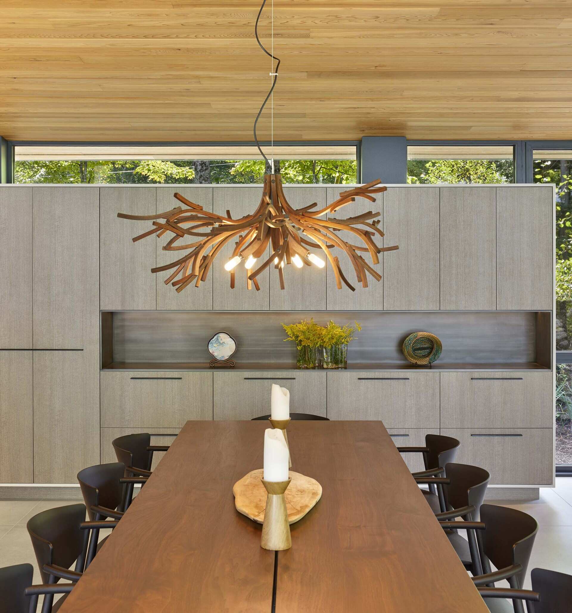 The dining area is located within the central great room and sits beside the lake-facing floor-to-ceiling windows. A custom solid walnut dining table is positioned within this space, accompanied by a chandelier made from recycled wood by Toronto makers The Brothers Dressler.