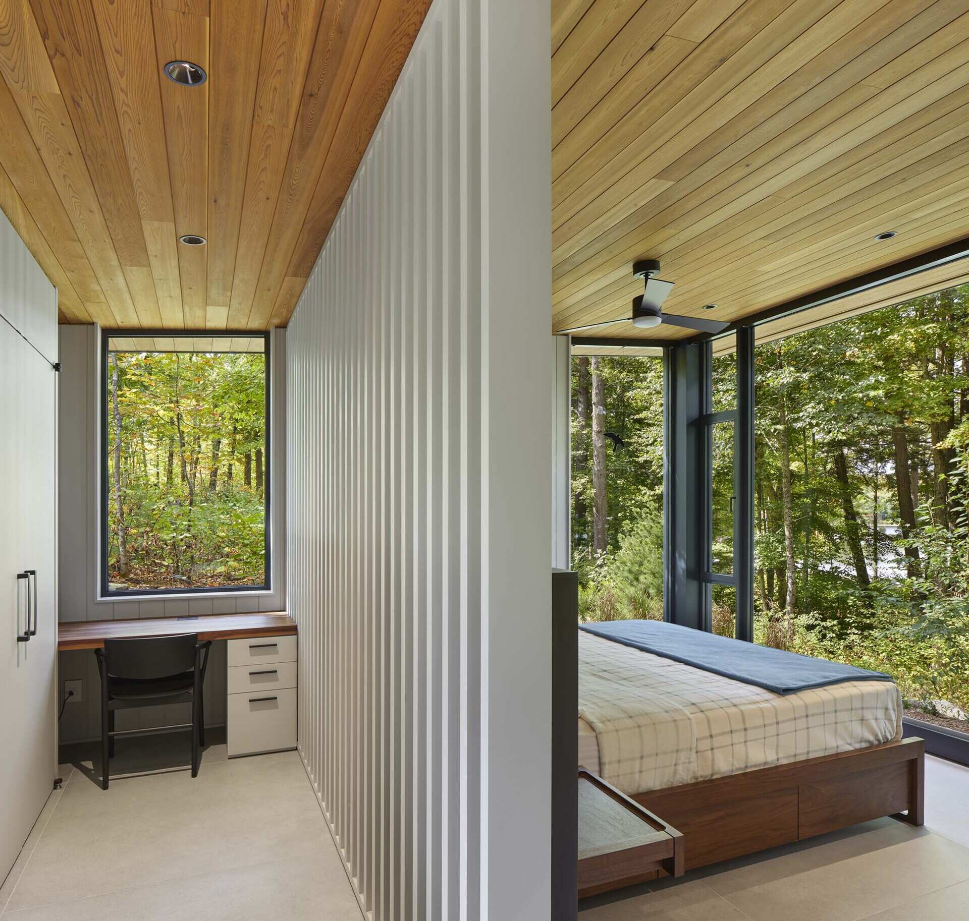In this primary bedroom suite, closets line the wall, a desk sits beneath a window, and a partition becomes a backdrop for the bed.
