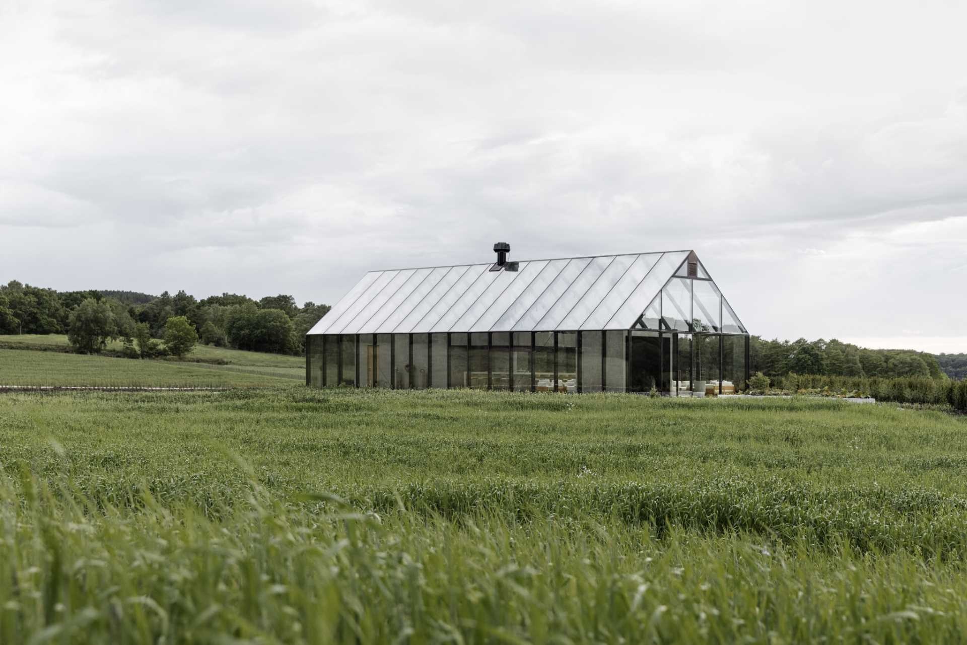 A winding meadow path leads through tall grasses toward a glass house restaurant with bold black frames standing in the middle of the field.