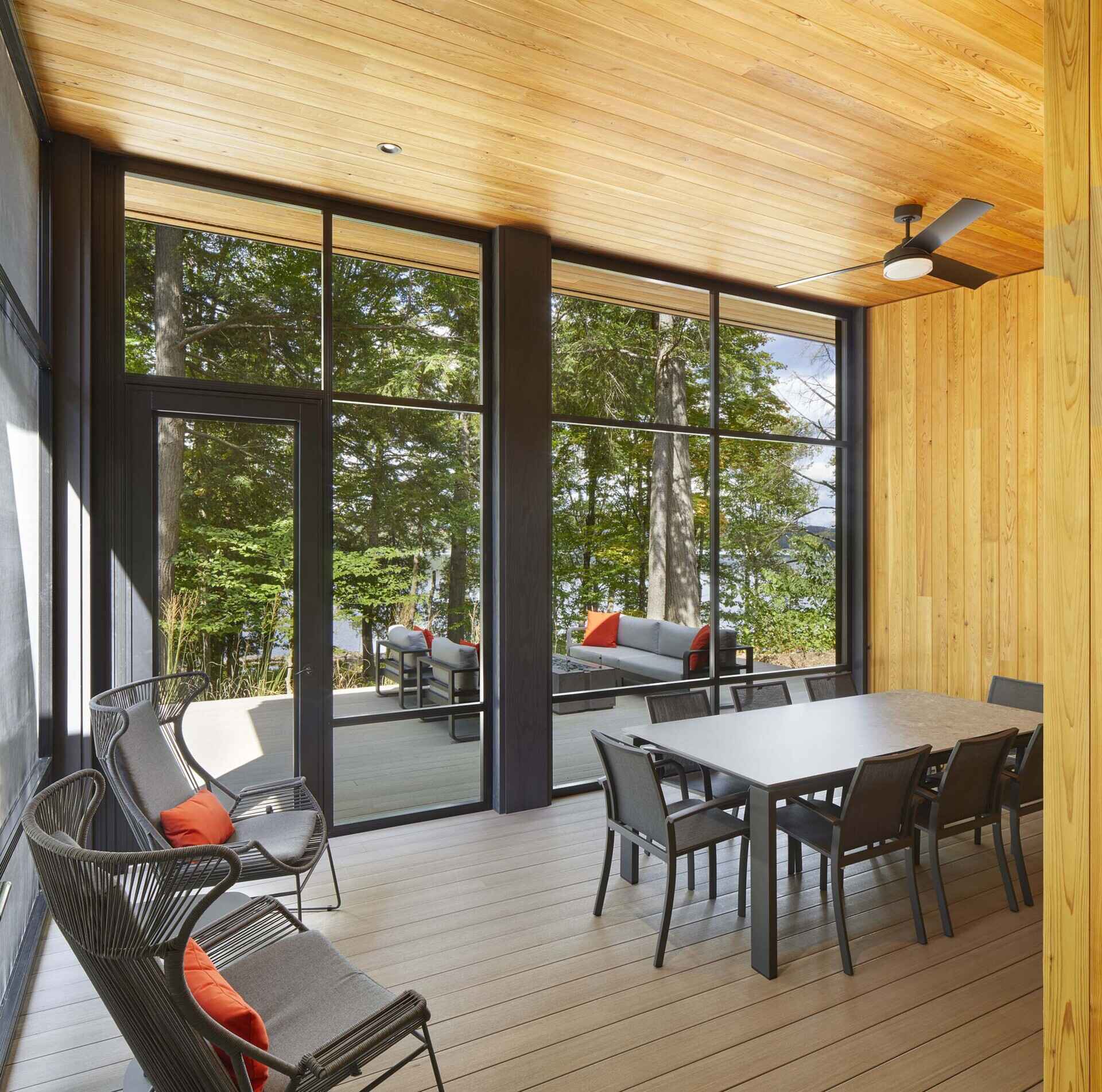 Located within the entry wing, the warm wood ceiling continues brings a cozy cabin-like feeling to this screened porch, while the large black frames open the space visually to the trees and water beyond, making the room feel connected to nature without giving up comfort.