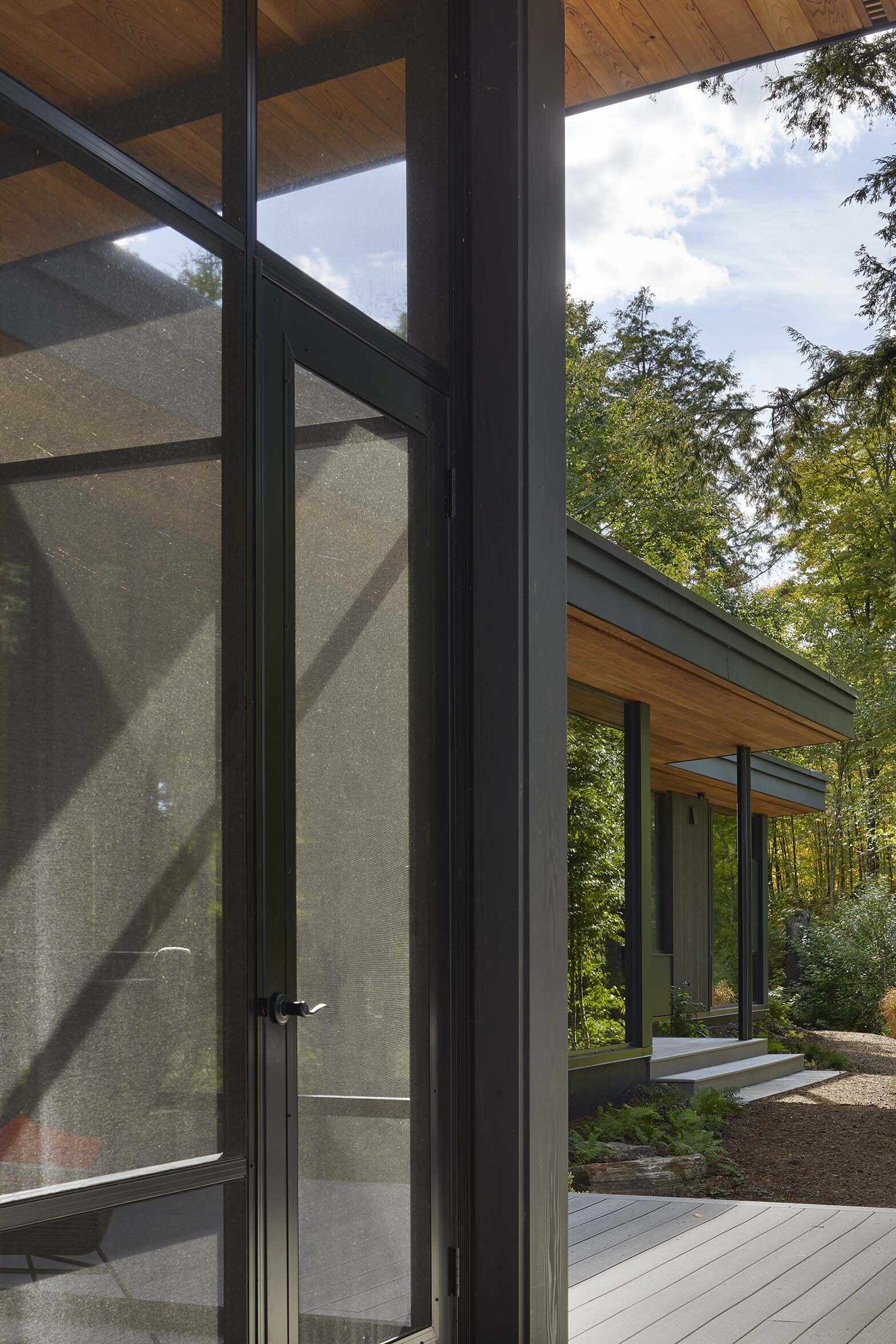 Located within the entry wing, the warm wood ceiling continues brings a cozy cabin-like feeling to this screened porch, while the large black frames open the space visually to the trees and water beyond, making the room feel connected to nature without giving up comfort.
