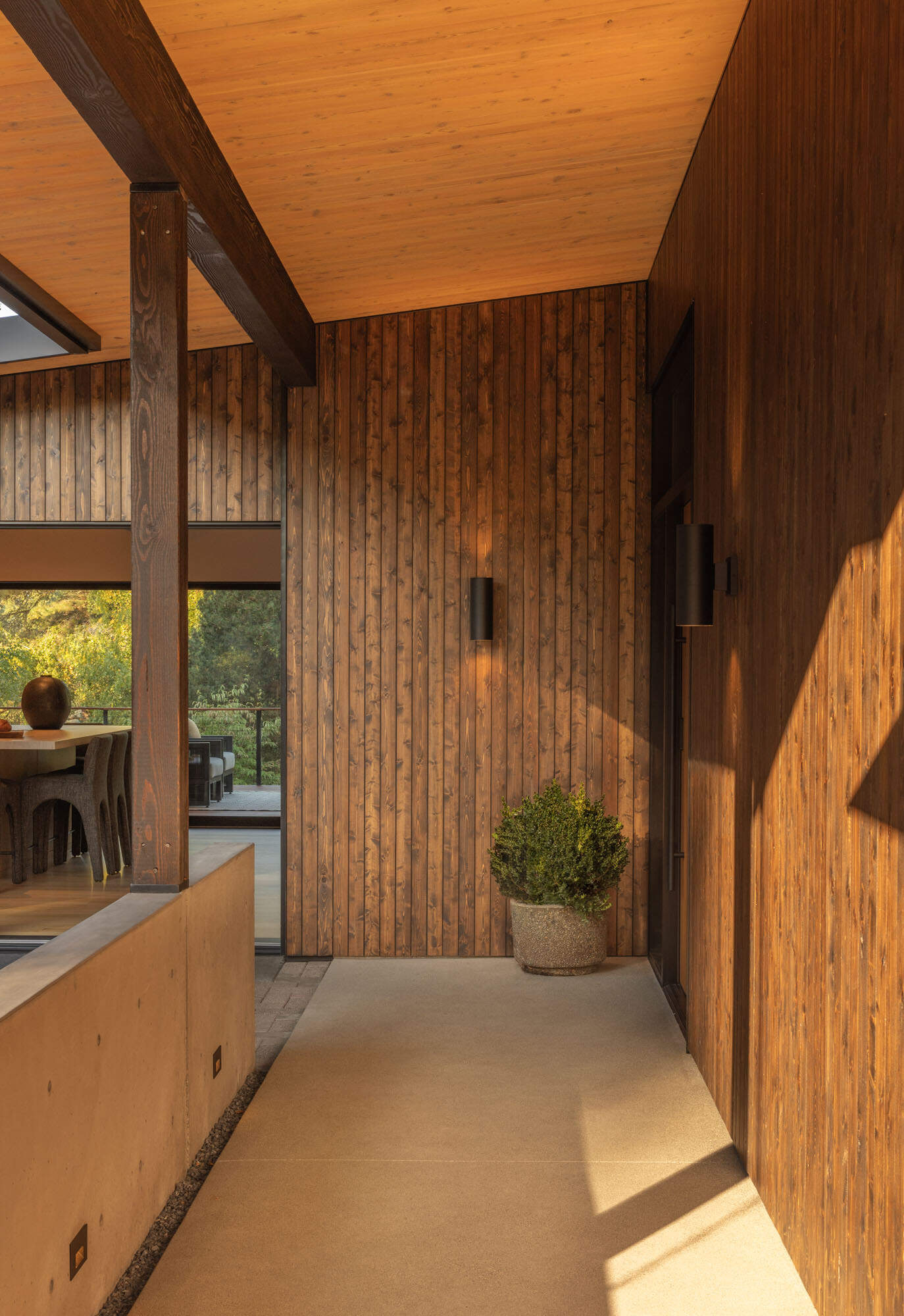 Vertical timber cladding wraps the facade, complementing the home's mid-century character, while black steel detailing adds crisp contrast around the door and windows. A sheltered porch provides a quiet transition from the courtyard into the home, balancing privacy with an open, welcoming feel.