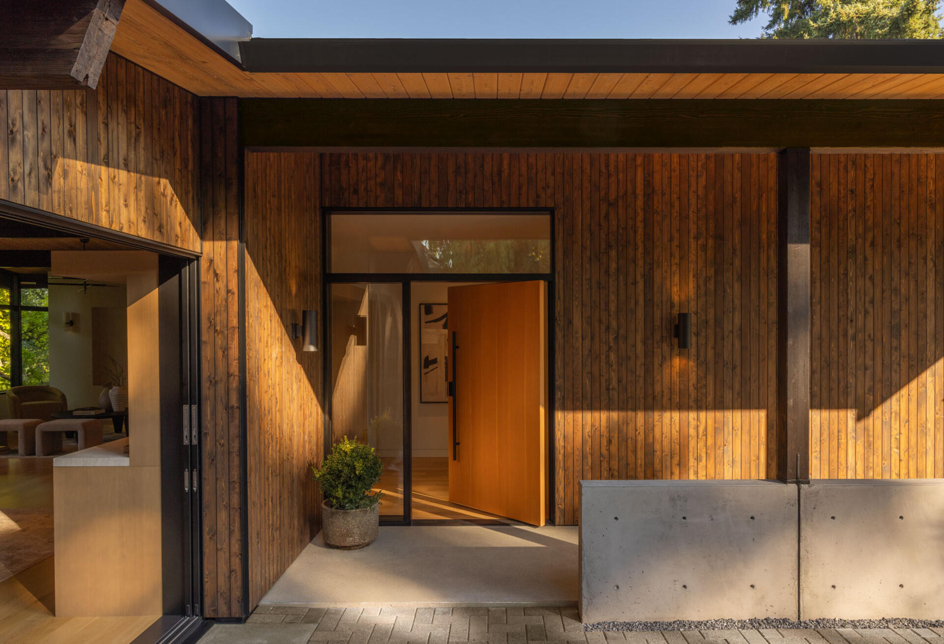 Set beneath a broad roof overhang, the oversized pivoting timber door is framed by full-height glazing that allows natural light to filter into the entryway while offering a glimpse of the warm interior beyond.