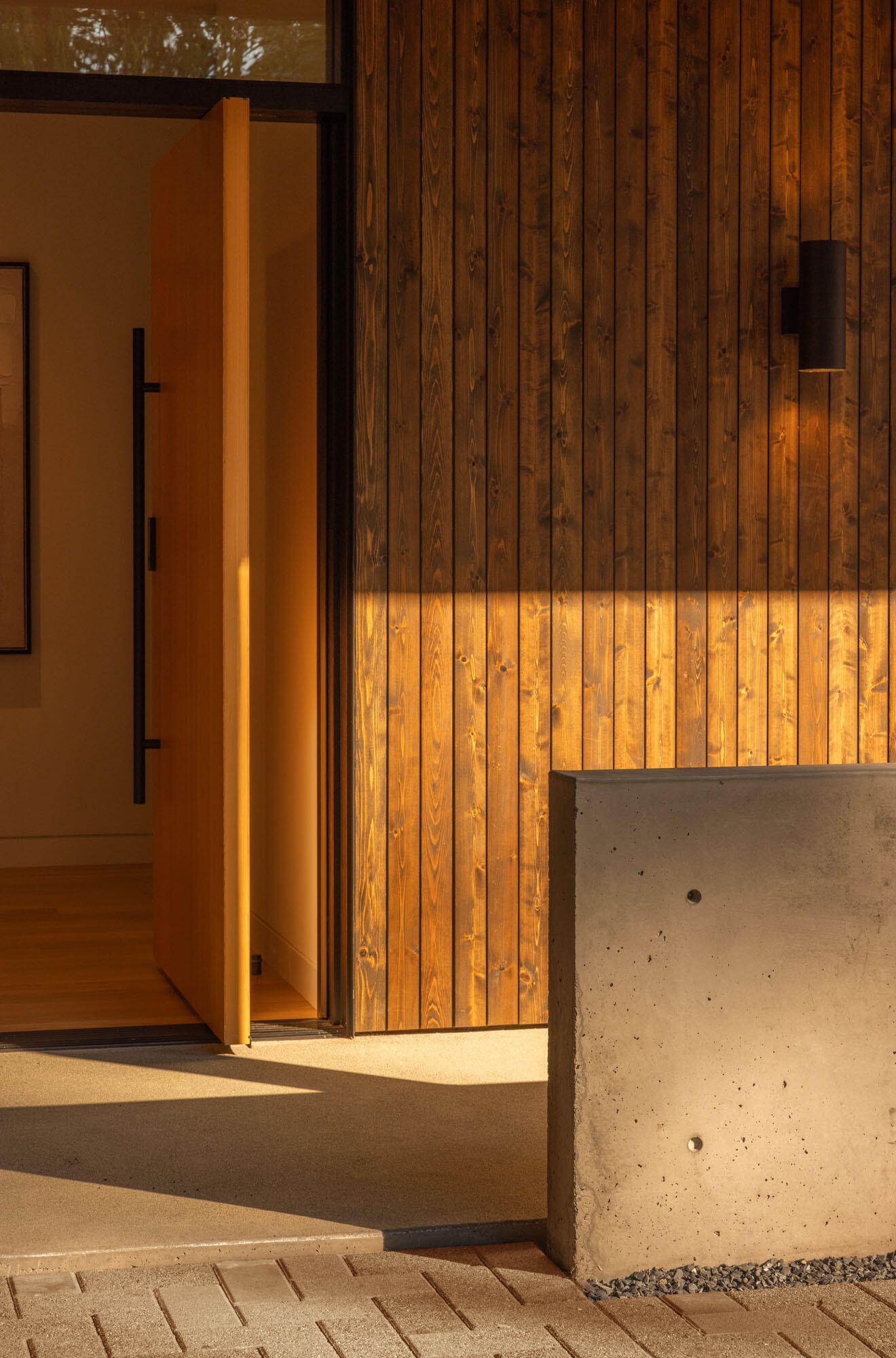 Set beneath a broad roof overhang, the oversized pivoting timber door is framed by full-height glazing that allows natural light to filter into the entryway while offering a glimpse of the warm interior beyond.