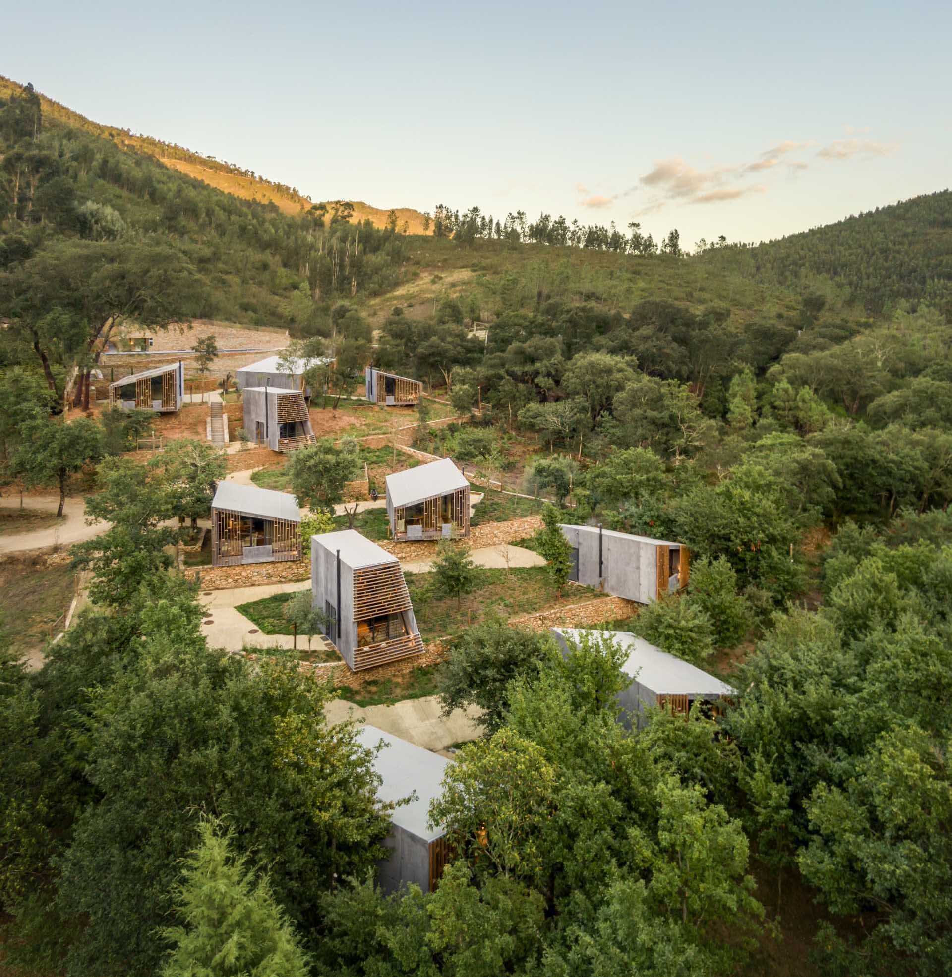 Eleven cabins in four unique designs are scattered across a Portuguese forest, preserving existing stone walls and mature trees. Their layout allows every home to enjoy open views of the surrounding woodland and river.