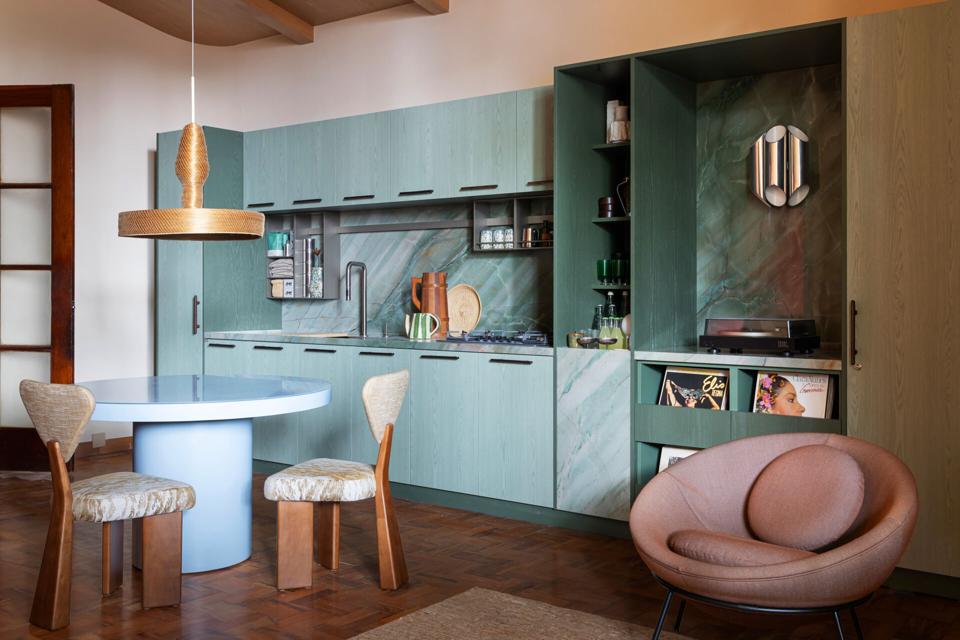 This kitchen and dining space combines green-toned cabinetry, quartzite surfaces, handcrafted lighting, and Brazilian design pieces in a stylish social setting.
