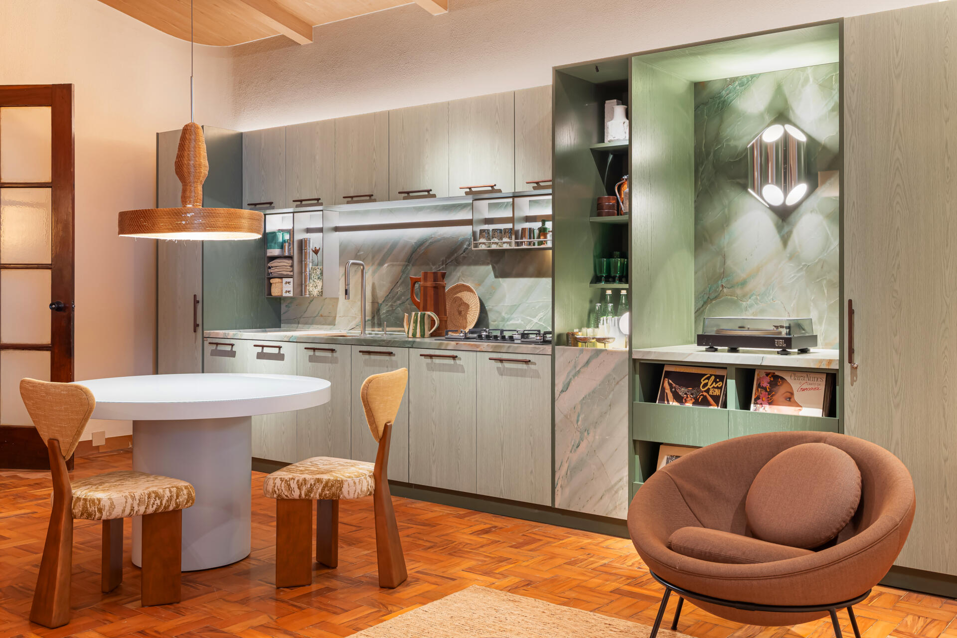 This kitchen and dining space combines green-toned cabinetry, quartzite surfaces, handcrafted lighting, and Brazilian design pieces in a stylish social setting.