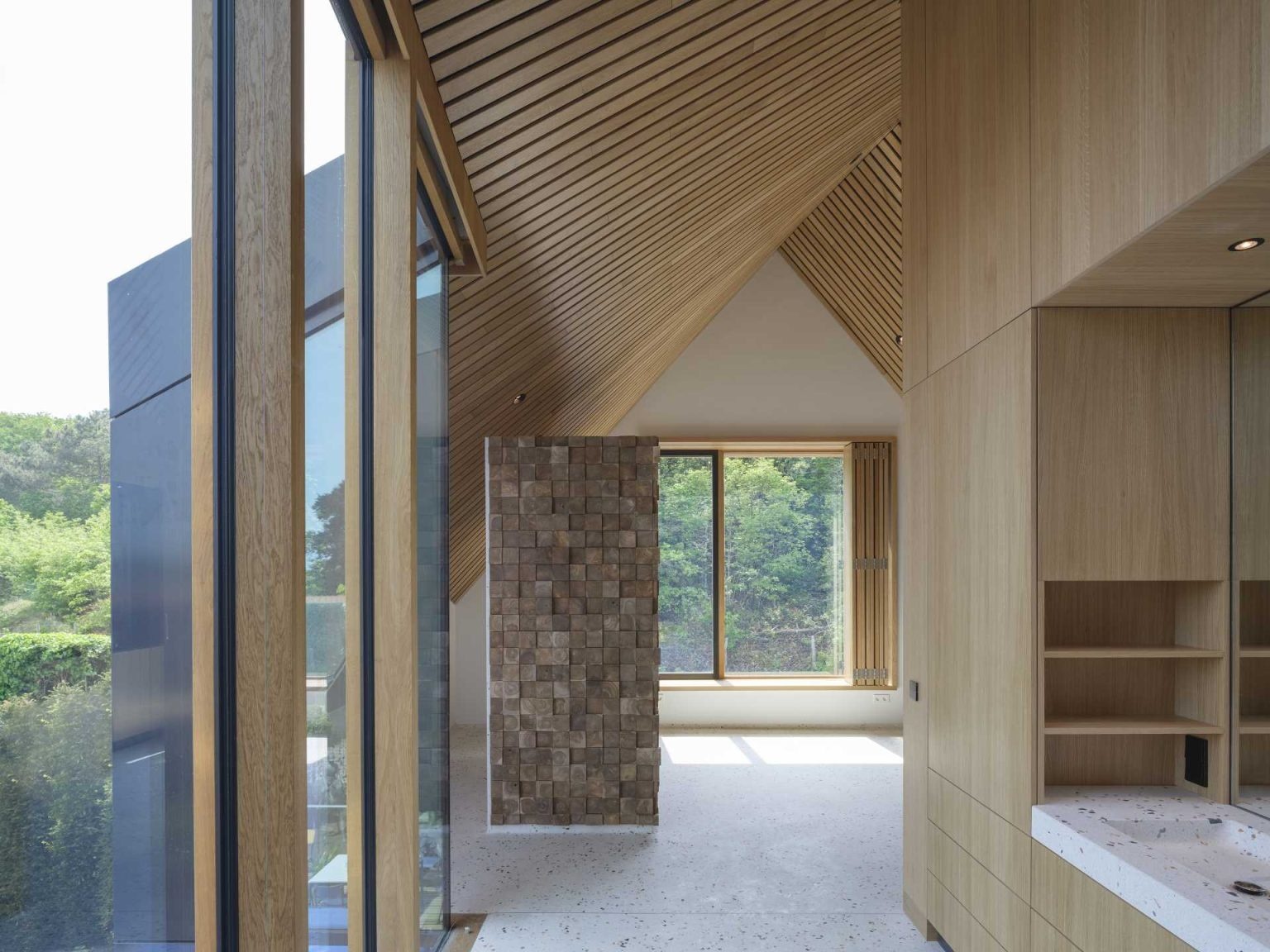 Inside, living spaces are organized around curtain wall-enclosed voids that draw natural light through the home. Wood-lined elements and framed countryside views create bright, open interiors that complement the dramatic exterior.