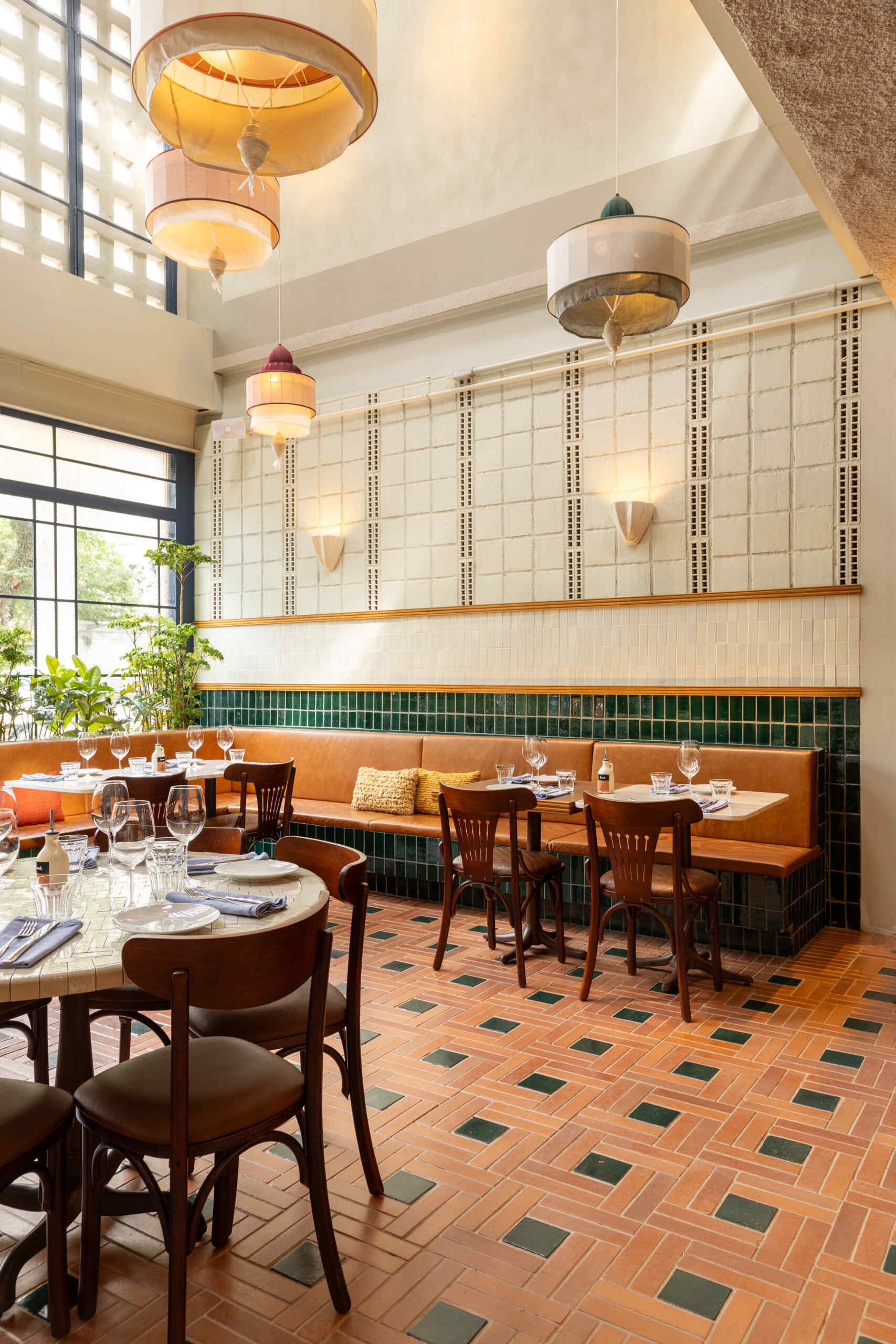 A double-height dining hall with terracotta geometric floors, green ceramic walls, concrete block relief patterns, and natural light from shed roofs creates a layered and welcoming restaurant interior.