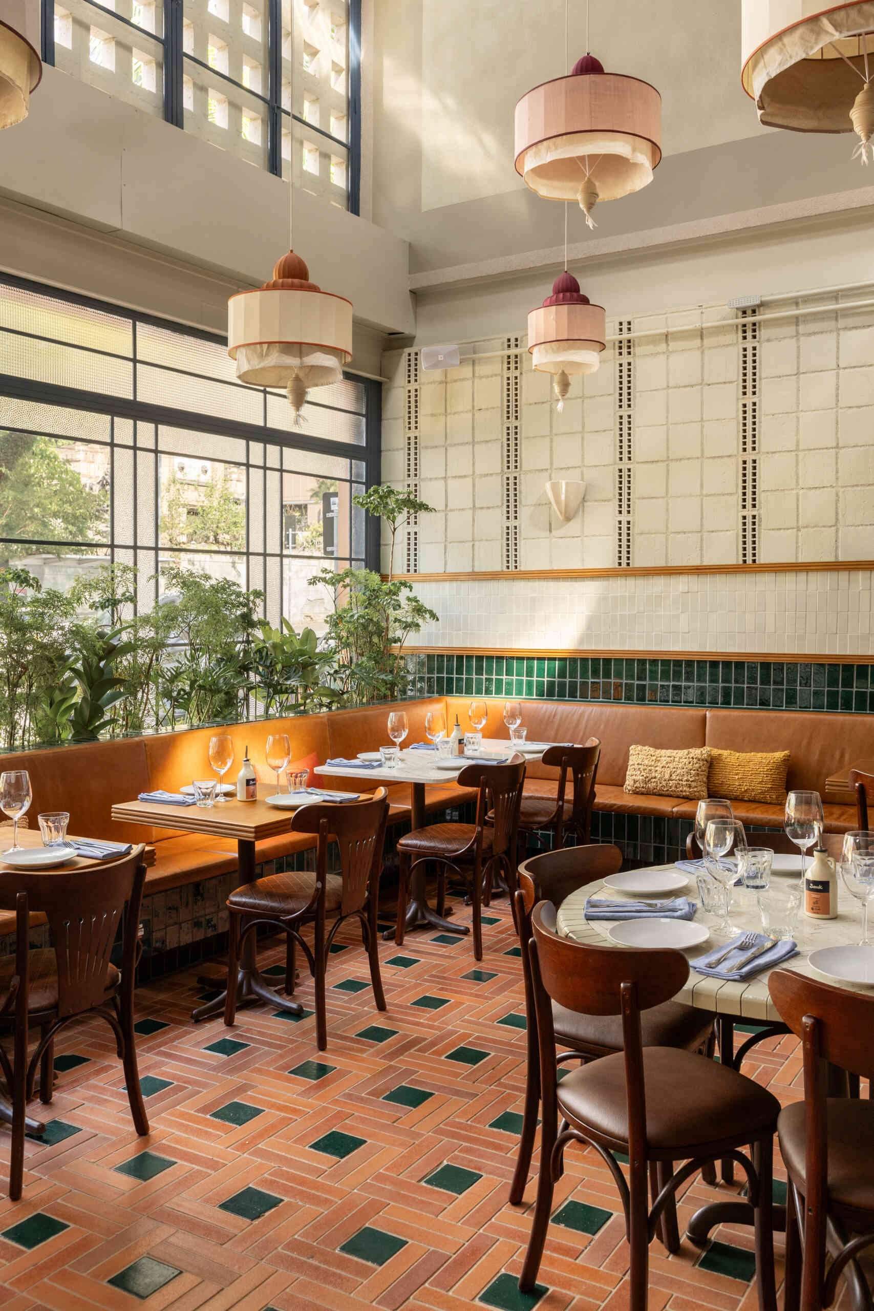 A double-height dining hall with terracotta geometric floors, green ceramic walls, concrete block relief patterns, and natural light from shed roofs creates a layered and welcoming restaurant interior.