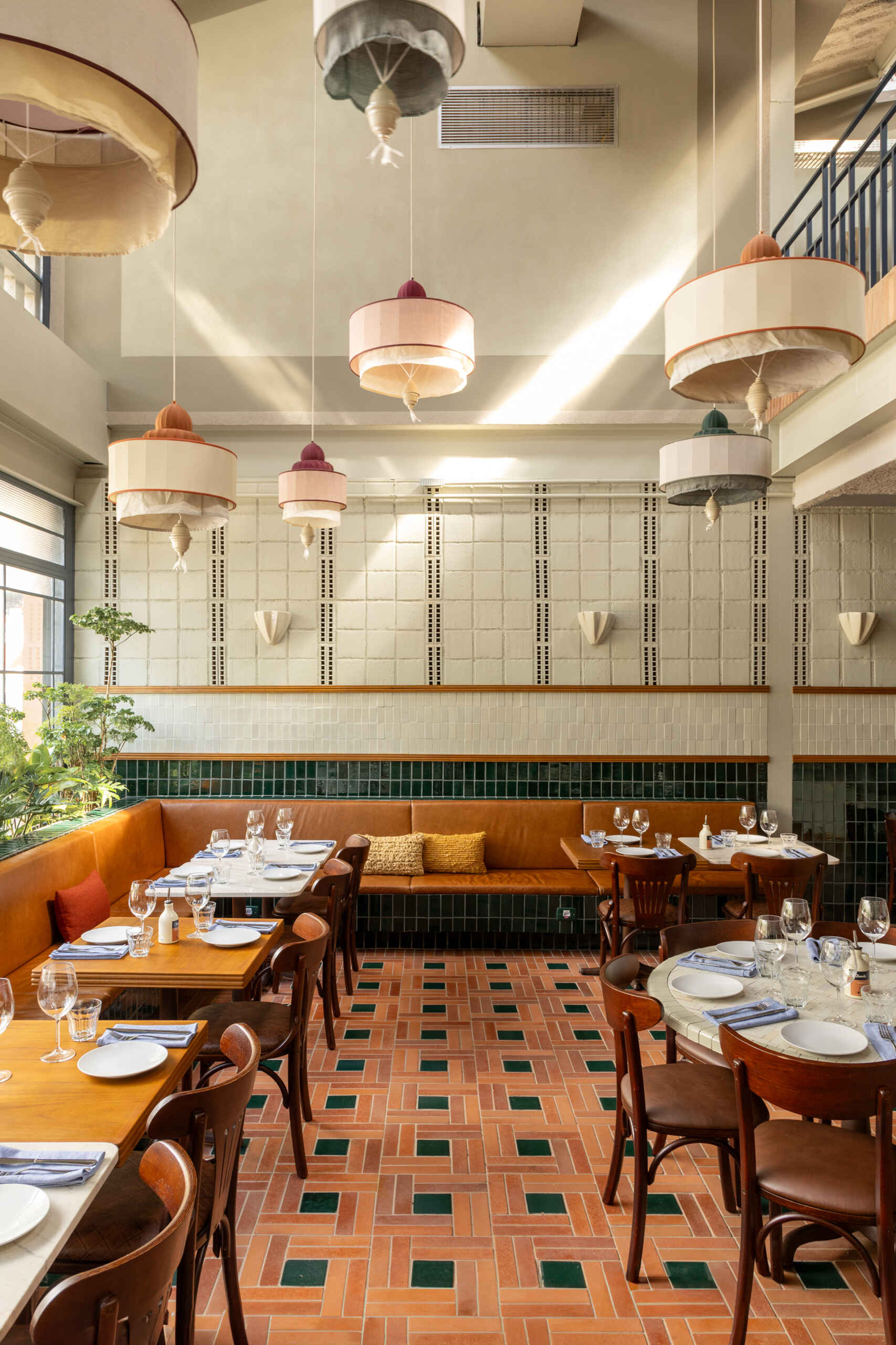 A double-height dining hall with terracotta geometric floors, green ceramic walls, concrete block relief patterns, and natural light from shed roofs creates a layered and welcoming restaurant interior.