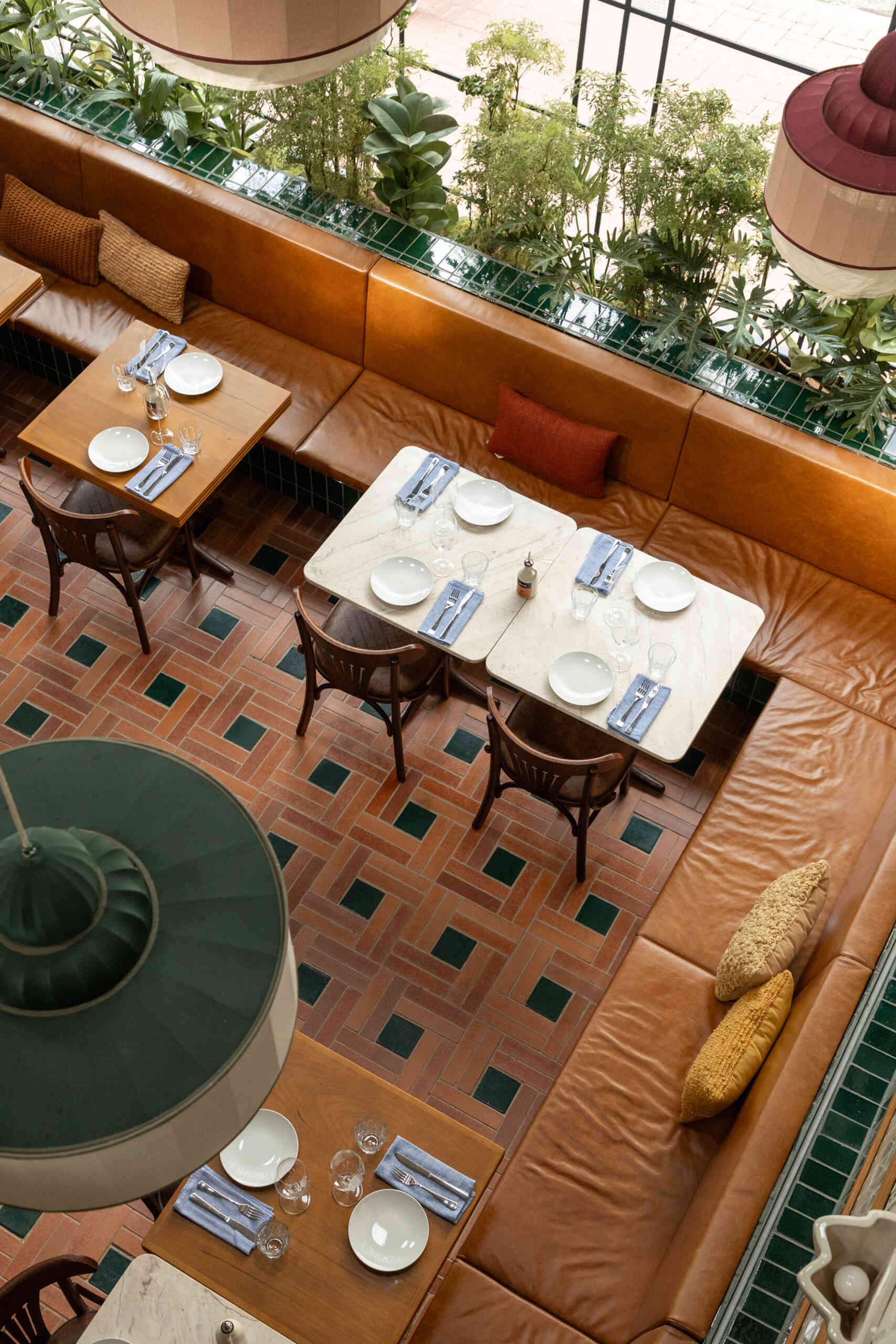 A double-height dining hall with terracotta geometric floors, green ceramic walls, concrete block relief patterns, and natural light from shed roofs creates a layered and welcoming restaurant interior.