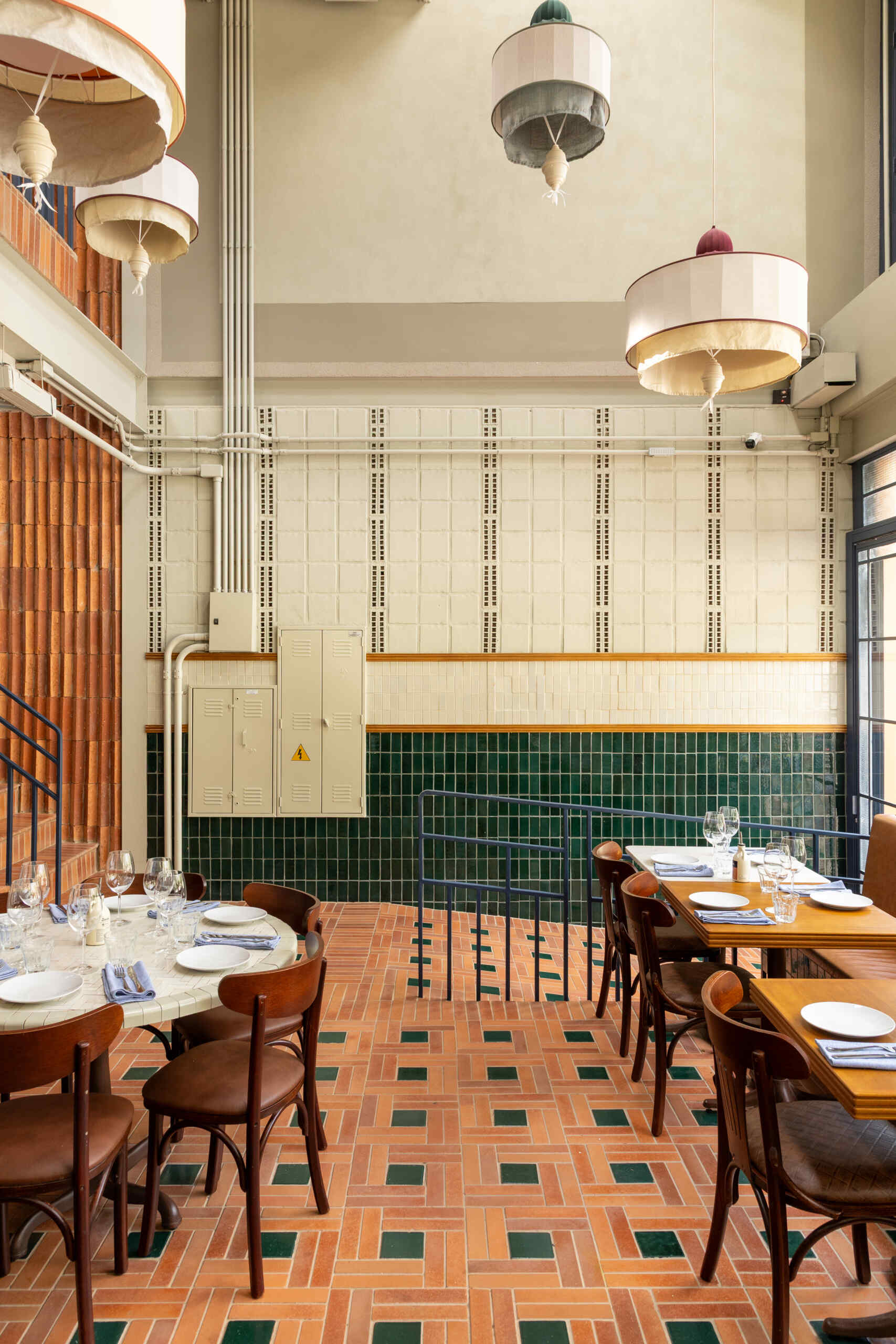 A double-height dining hall with terracotta geometric floors, green ceramic walls, concrete block relief patterns, and natural light from shed roofs creates a layered and welcoming restaurant interior.