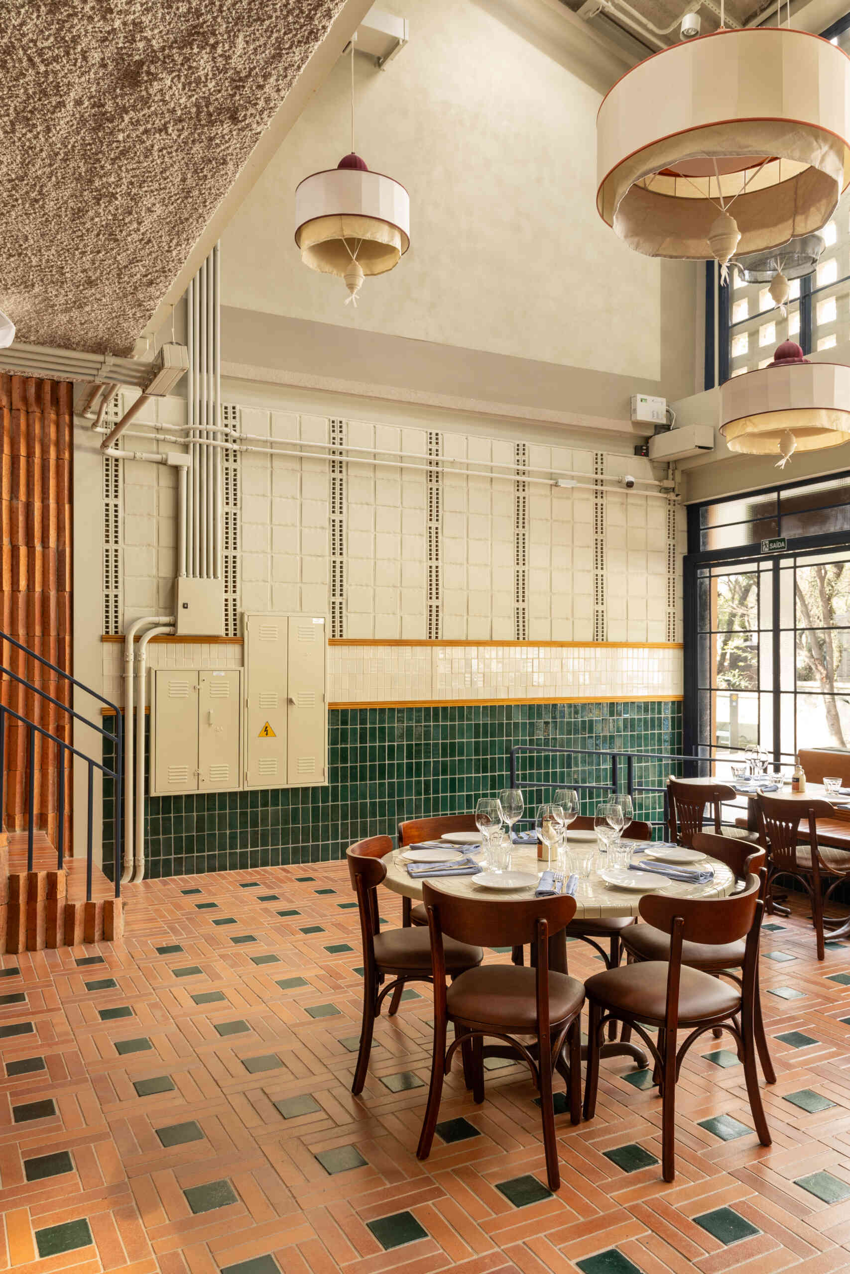A double-height dining hall with terracotta geometric floors, green ceramic walls, concrete block relief patterns, and natural light from shed roofs creates a layered and welcoming restaurant interior.