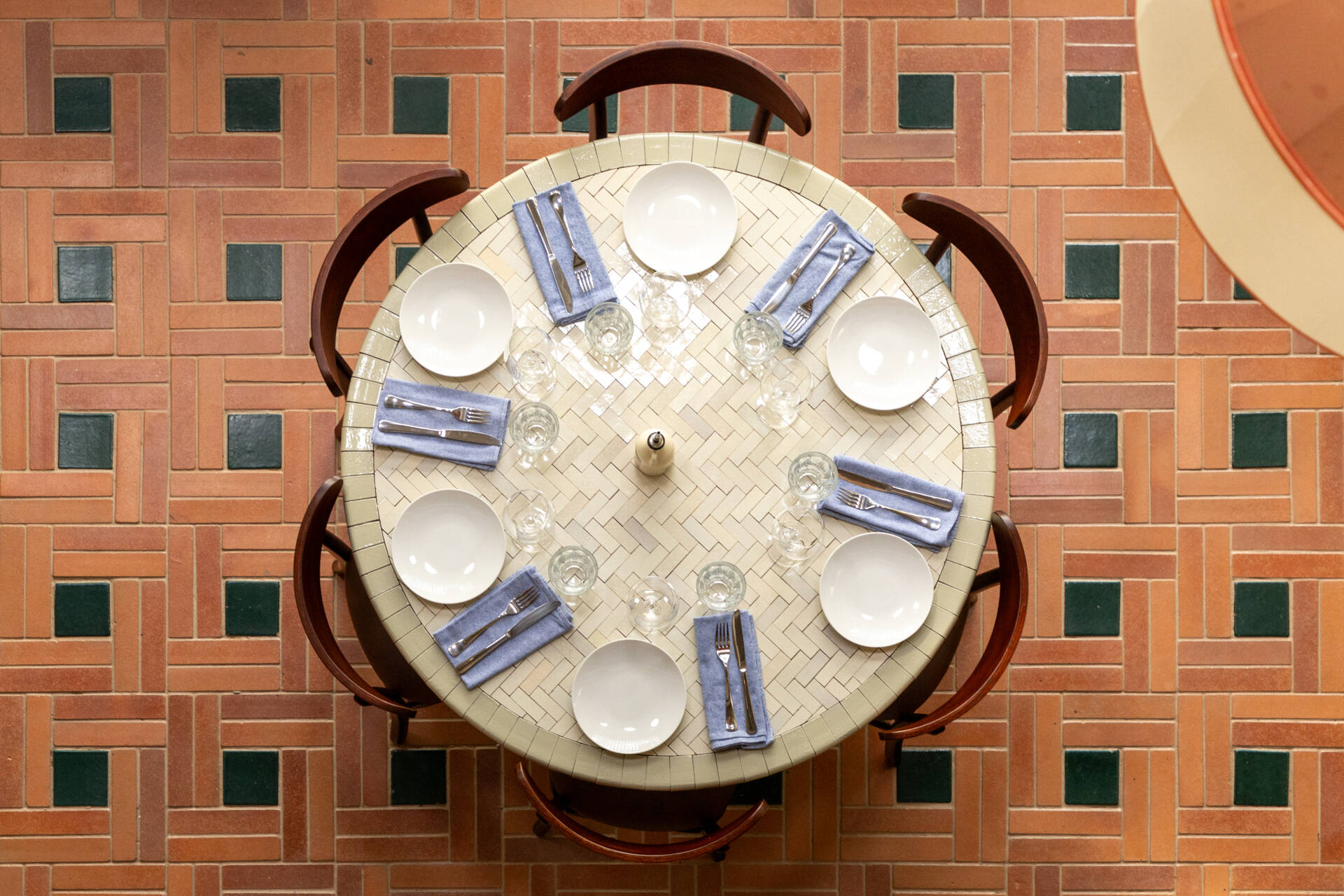 A double-height dining hall with terracotta geometric floors, green ceramic walls, concrete block relief patterns, and natural light from shed roofs creates a layered and welcoming restaurant interior.