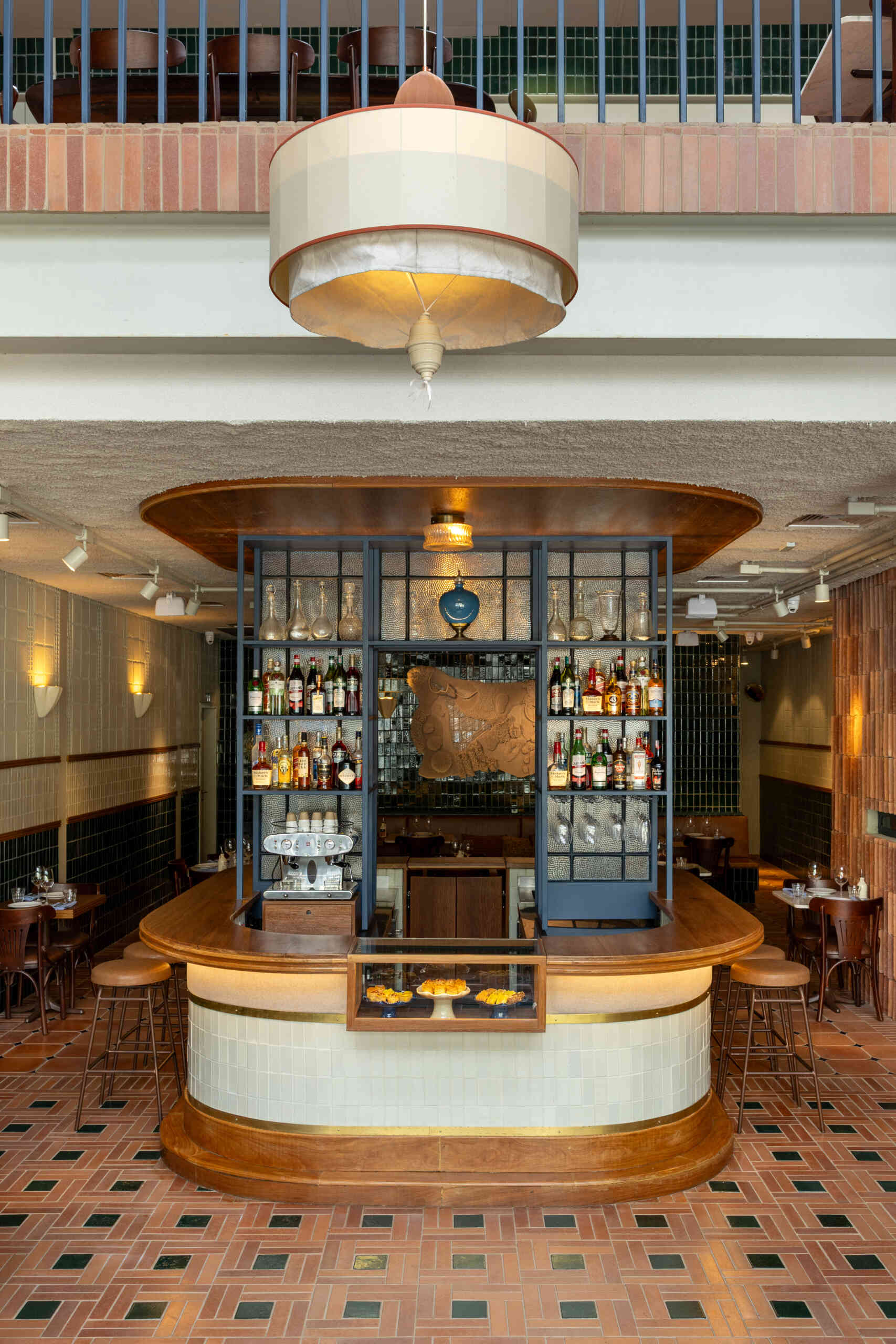 The rounded central bar features ceramic cladding, brass detailing, warm indirect lighting, and a blue iron display structure that acts like a portal framing the restaurant’s beverage collection.