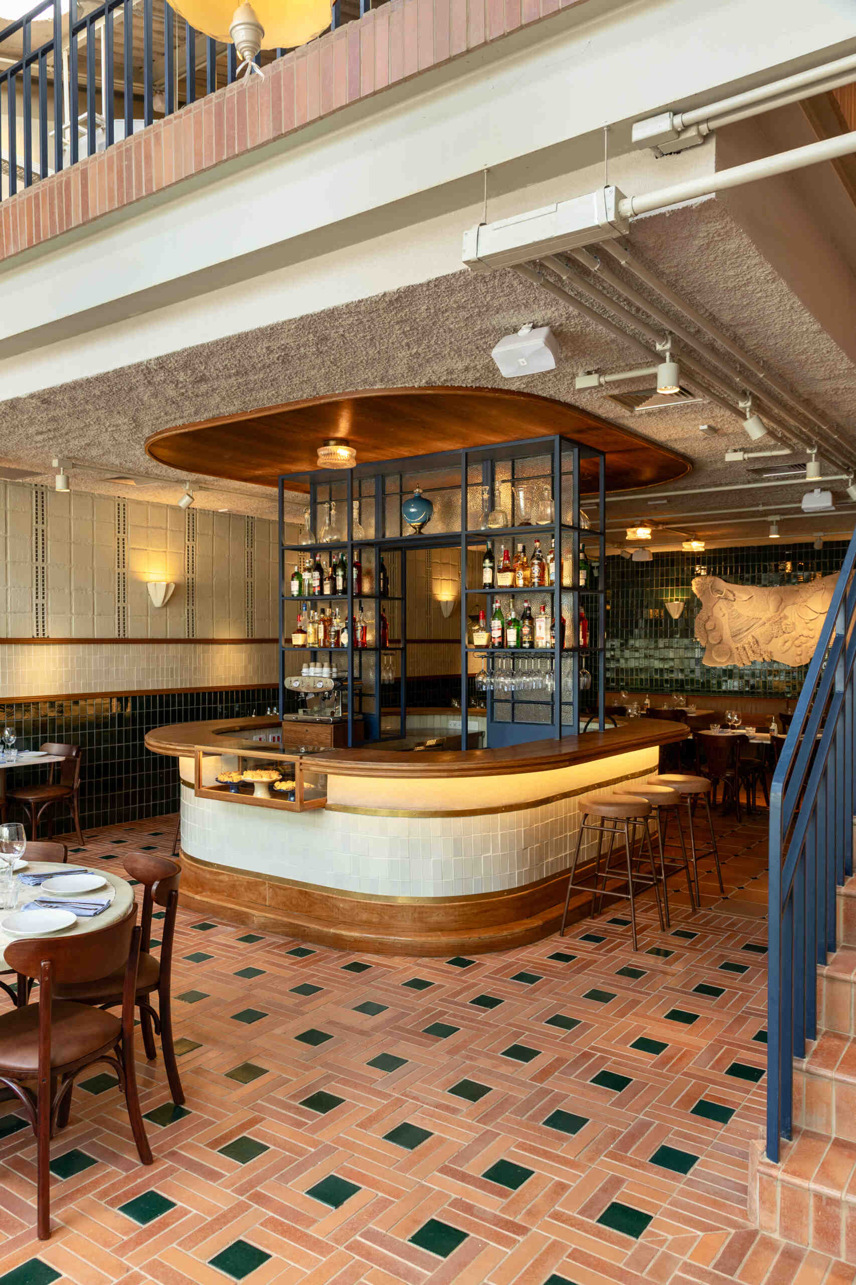 The rounded central bar features ceramic cladding, brass detailing, warm indirect lighting, and a blue iron display structure that acts like a portal framing the restaurant’s beverage collection.