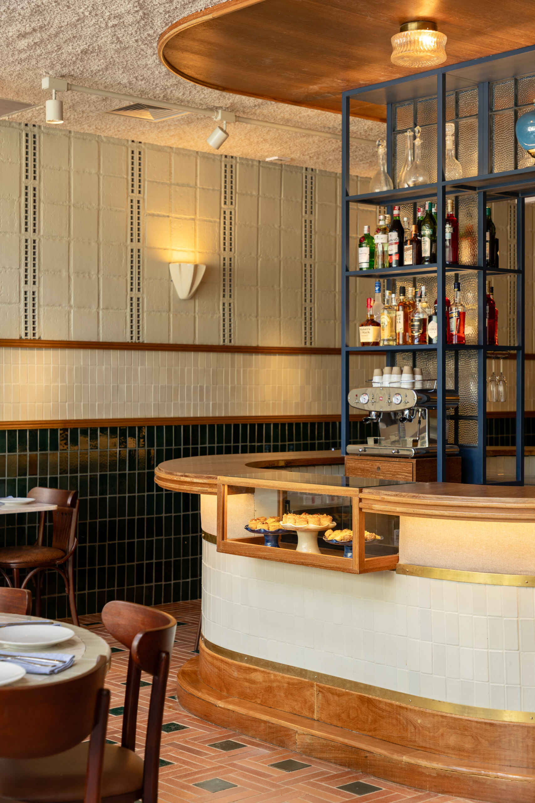 The rounded central bar features ceramic cladding, brass detailing, warm indirect lighting, and a blue iron display structure that acts like a portal framing the restaurant’s beverage collection.