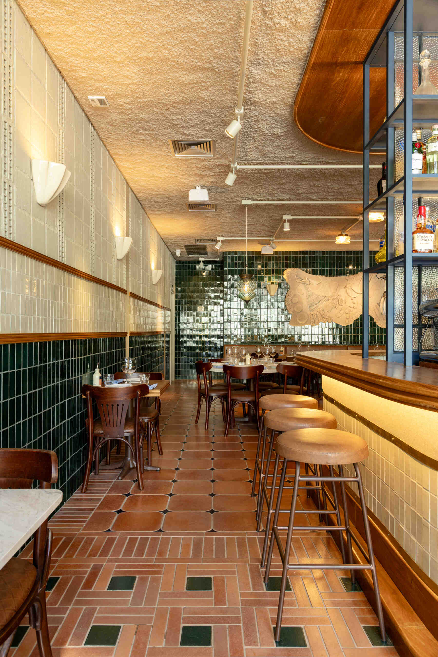 A double-height dining hall with terracotta geometric floors, green ceramic walls, concrete block relief patterns, and natural light from shed roofs creates a layered and welcoming restaurant interior.