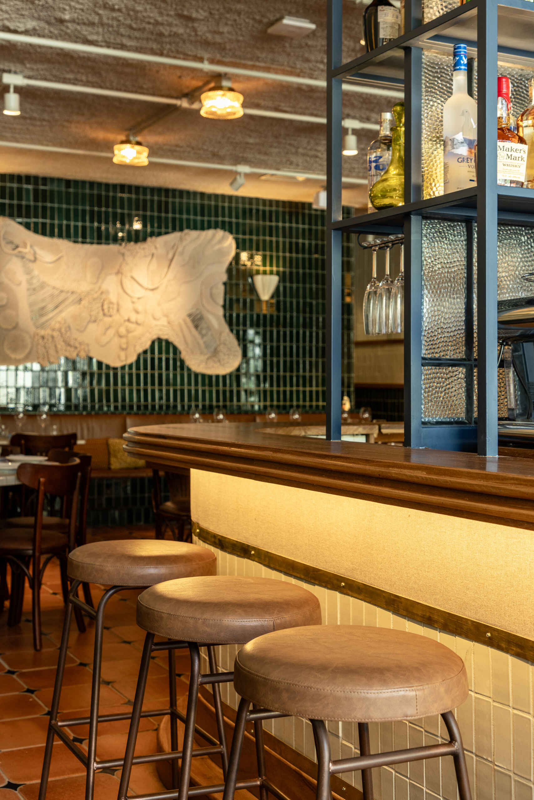 The rounded central bar features ceramic cladding, brass detailing, warm indirect lighting, and a blue iron display structure that acts like a portal framing the restaurant’s beverage collection.