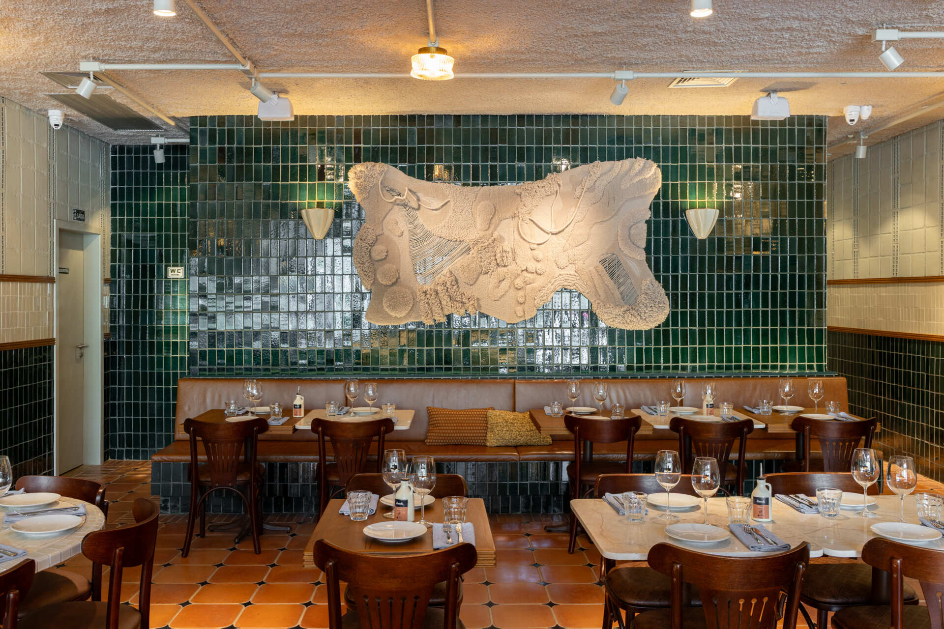 A double-height dining hall with terracotta geometric floors, green ceramic walls, concrete block relief patterns, and natural light from shed roofs creates a layered and welcoming restaurant interior.