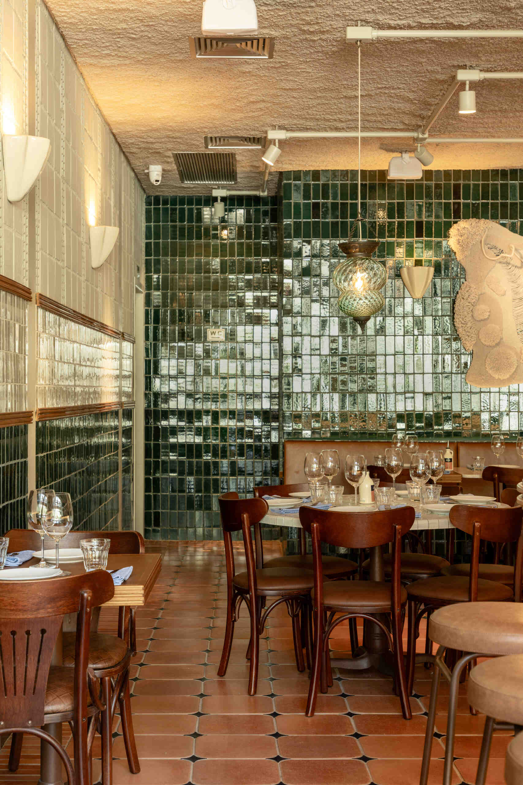A double-height dining hall with terracotta geometric floors, green ceramic walls, concrete block relief patterns, and natural light from shed roofs creates a layered and welcoming restaurant interior.
