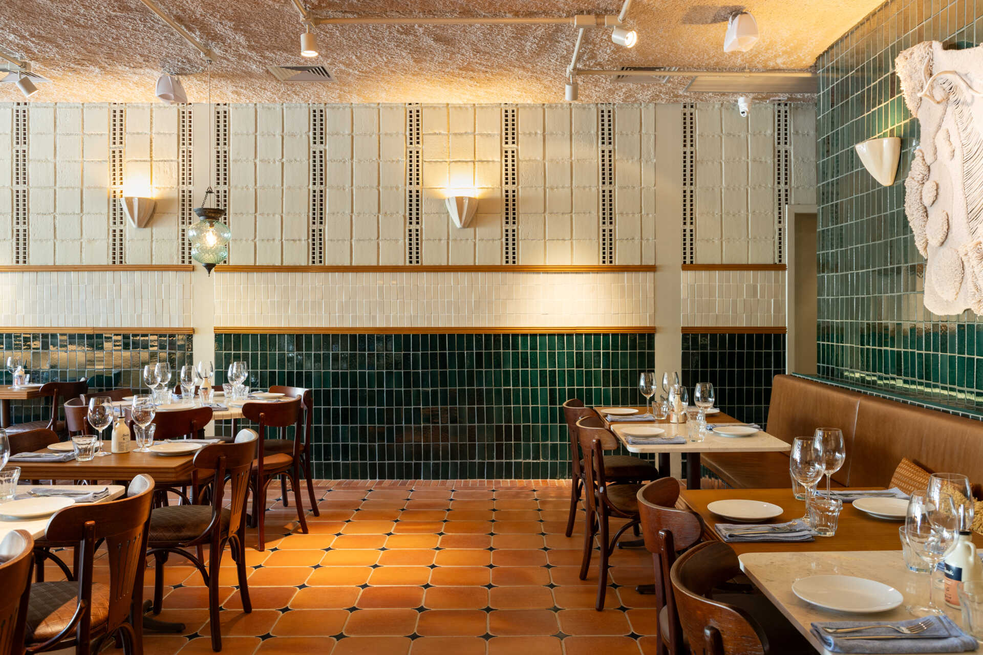 A double-height dining hall with terracotta geometric floors, green ceramic walls, concrete block relief patterns, and natural light from shed roofs creates a layered and welcoming restaurant interior.