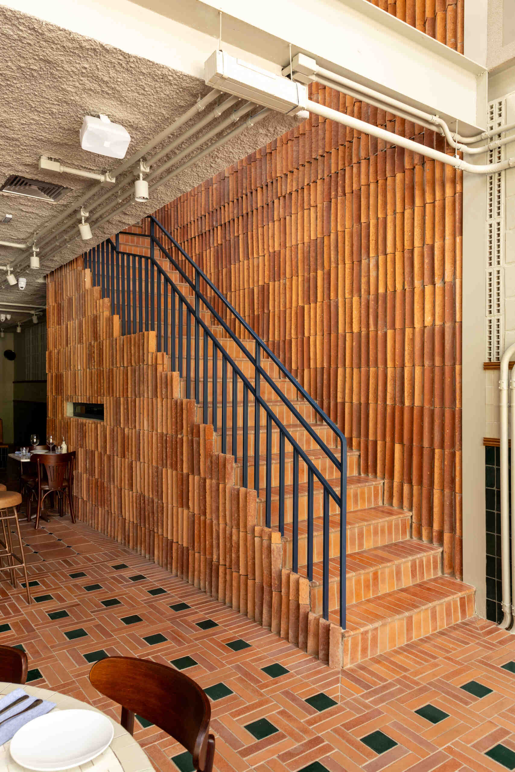 Terracotta stair treads, a blue iron railing, and triangular ceramic tile pieces create a sculptural staircase where light enhances the three-dimensional surface pattern.