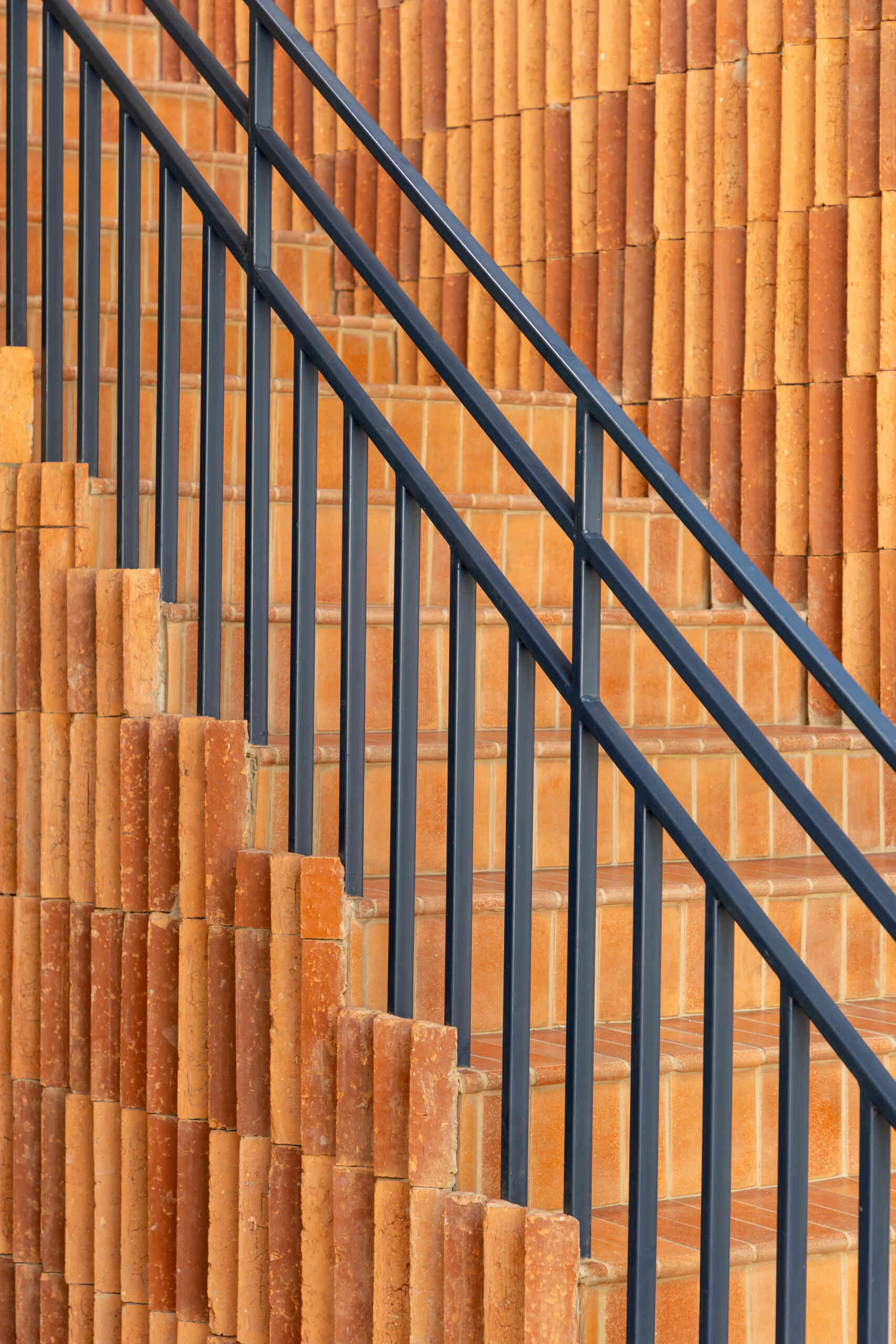 Terracotta stair treads, a blue iron railing, and triangular ceramic tile pieces create a sculptural staircase where light enhances the three-dimensional surface pattern.