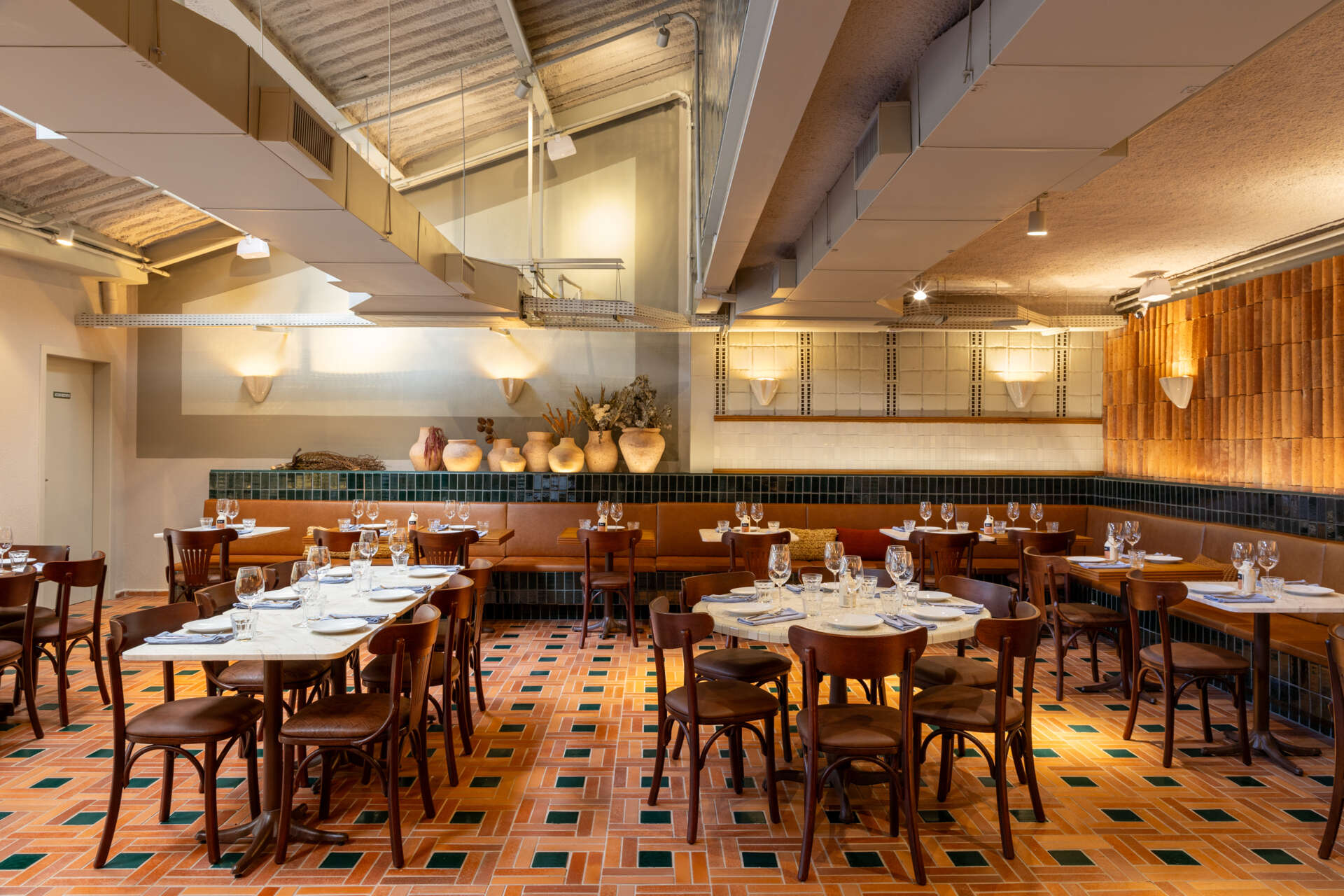 The mezzanine is filled with filtered natural light and overlooks the double-height dining hall, with terracotta, green ceramics, and blue iron details continuing the material language upstairs.