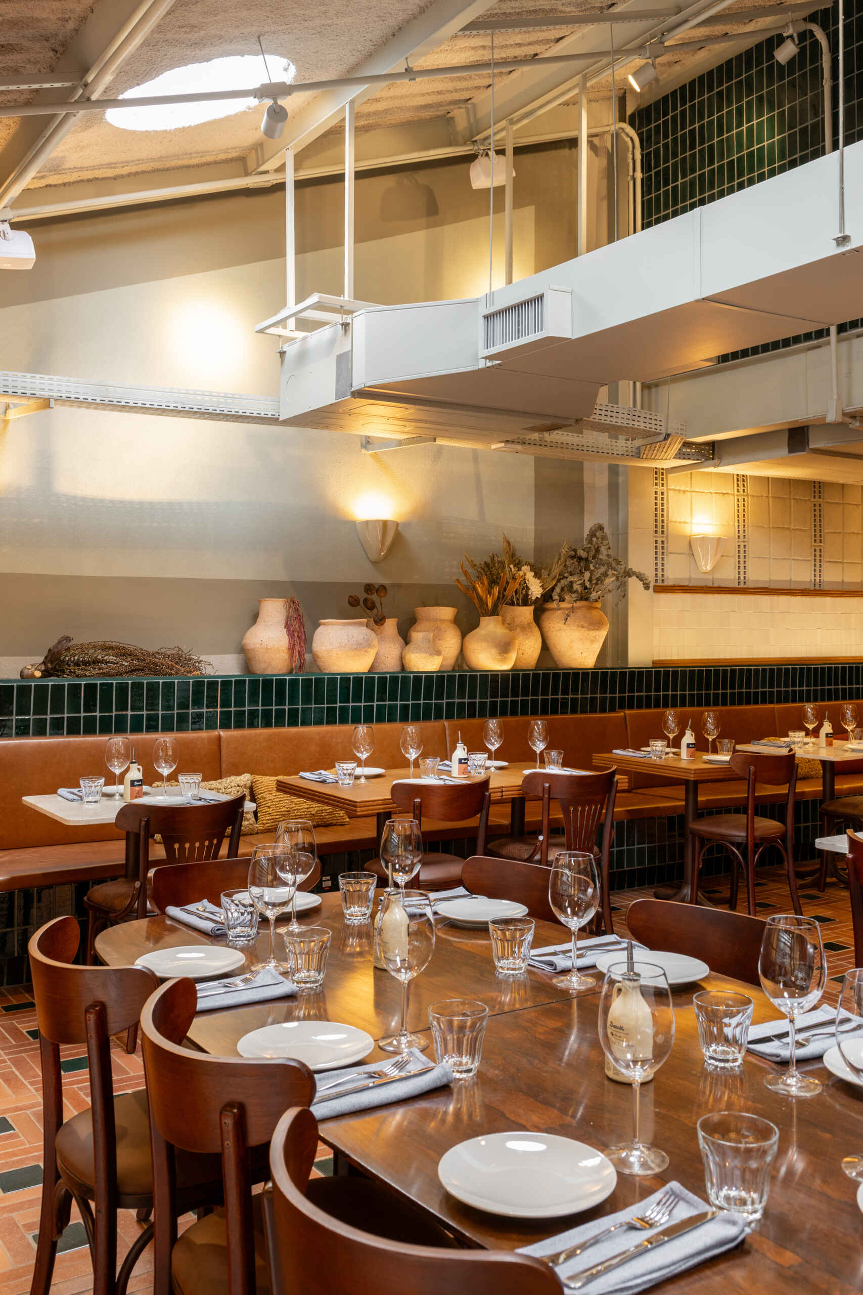 The mezzanine is filled with filtered natural light and overlooks the double-height dining hall, with terracotta, green ceramics, and blue iron details continuing the material language upstairs.