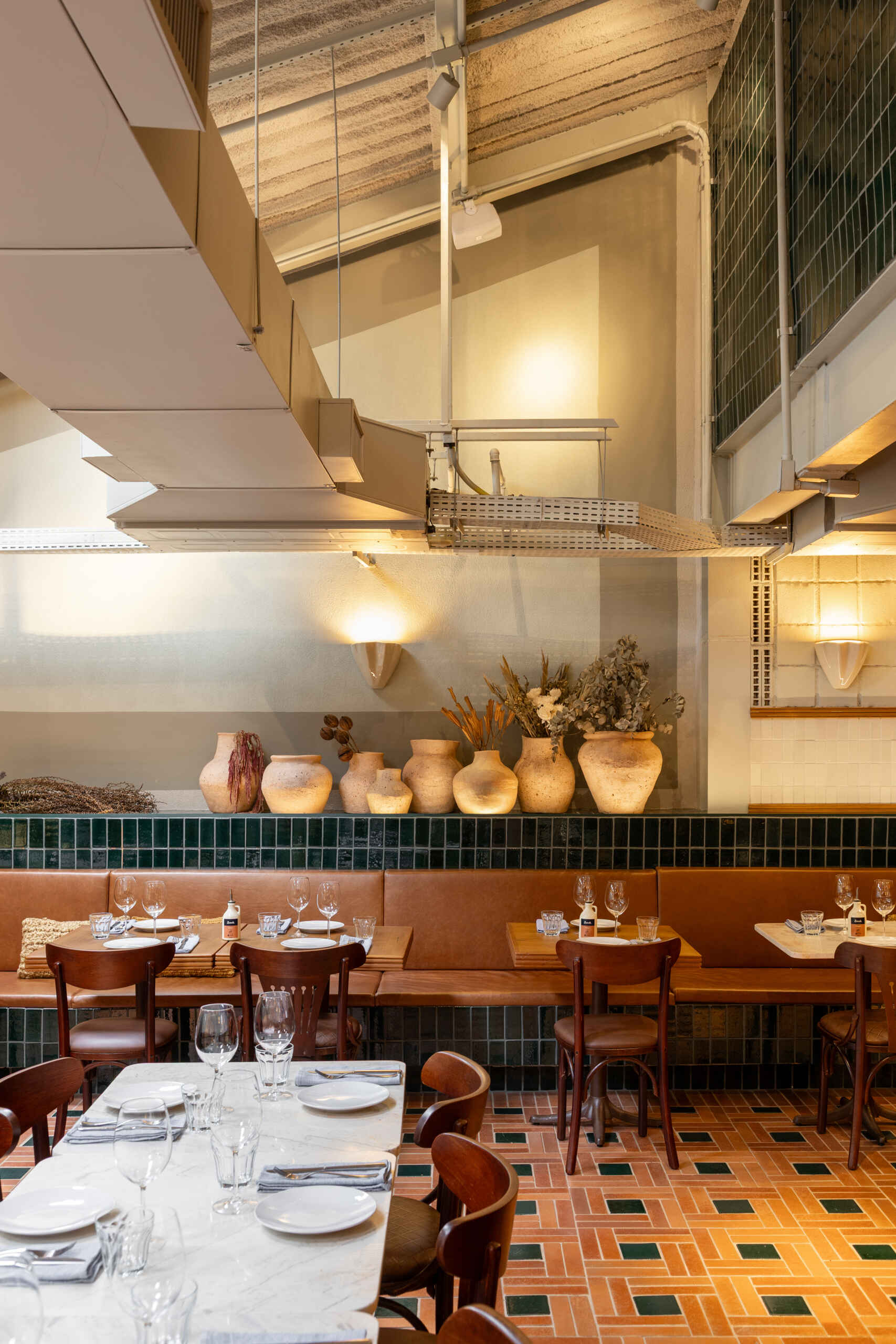 The mezzanine is filled with filtered natural light and overlooks the double-height dining hall, with terracotta, green ceramics, and blue iron details continuing the material language upstairs.