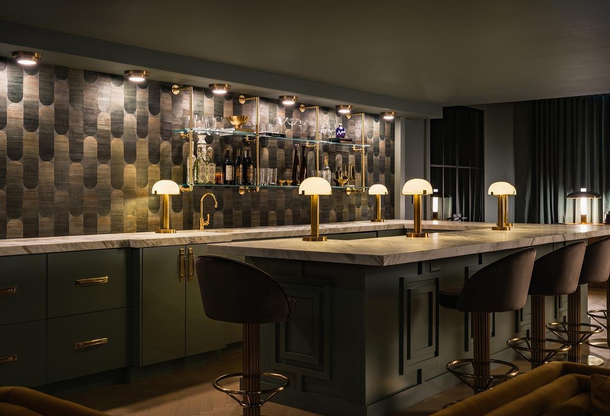 This speakeasy-inspired home whiskey lounge features rich green cabinetry, South African marble, brass accents, velvet seating, and vintage-inspired details.
