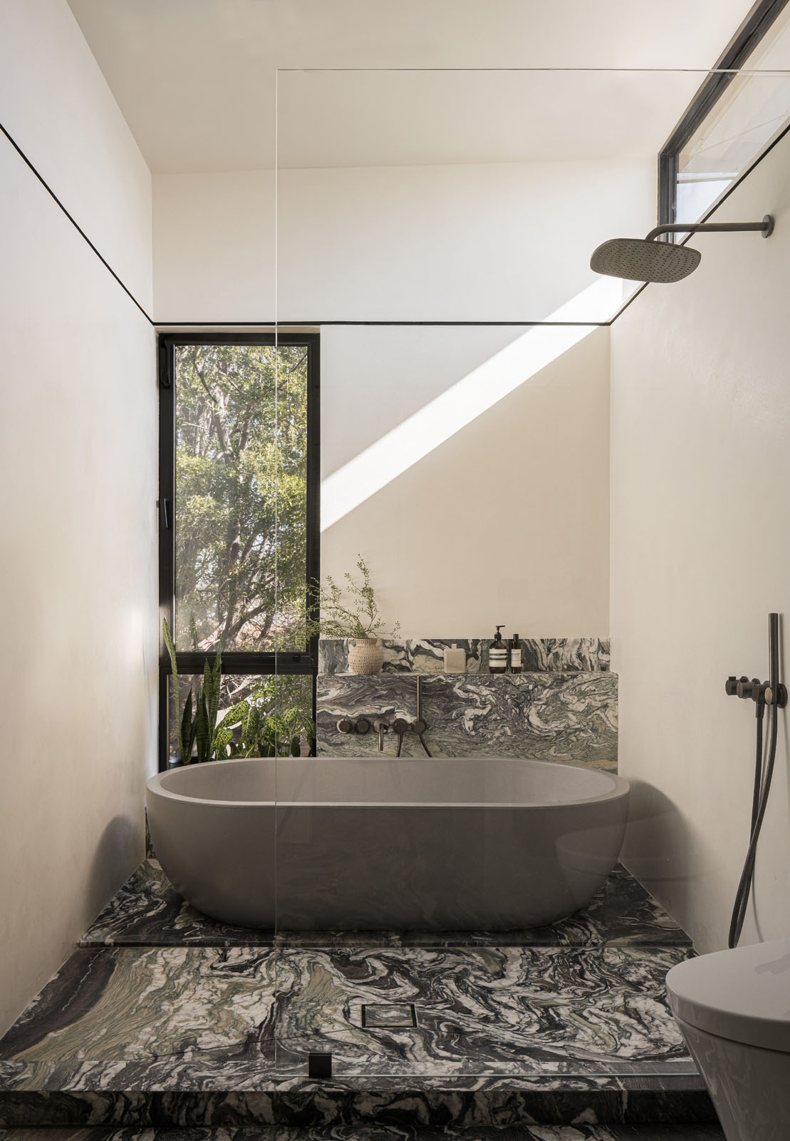 Bold veined stone, sculptural vanities, and a freestanding bath beside a tree-filled window give this primary bathroom the feel of a luxurious spa.