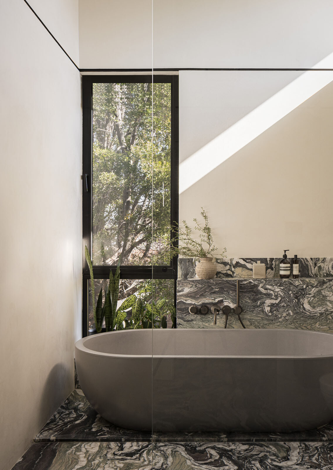 Bold veined stone, sculptural vanities, and a freestanding bath beside a tree-filled window give this primary bathroom the feel of a luxurious spa.