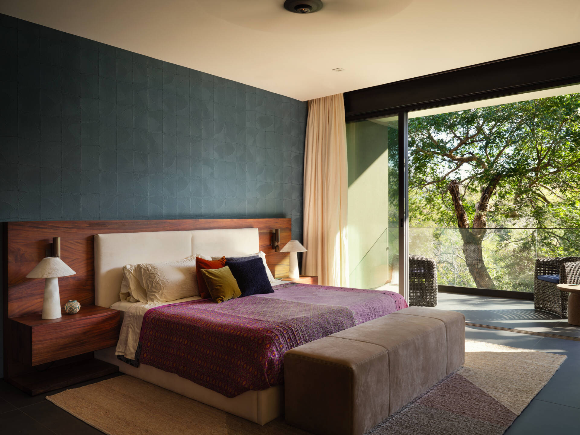 A textured blue feature wall brings depth to this bedroom, complemented by a wide timber headboard and softly upholstered bed. Plum bedding and jewel-toned cushions introduce color, while the restrained palette keeps the space feeling calm and inviting.