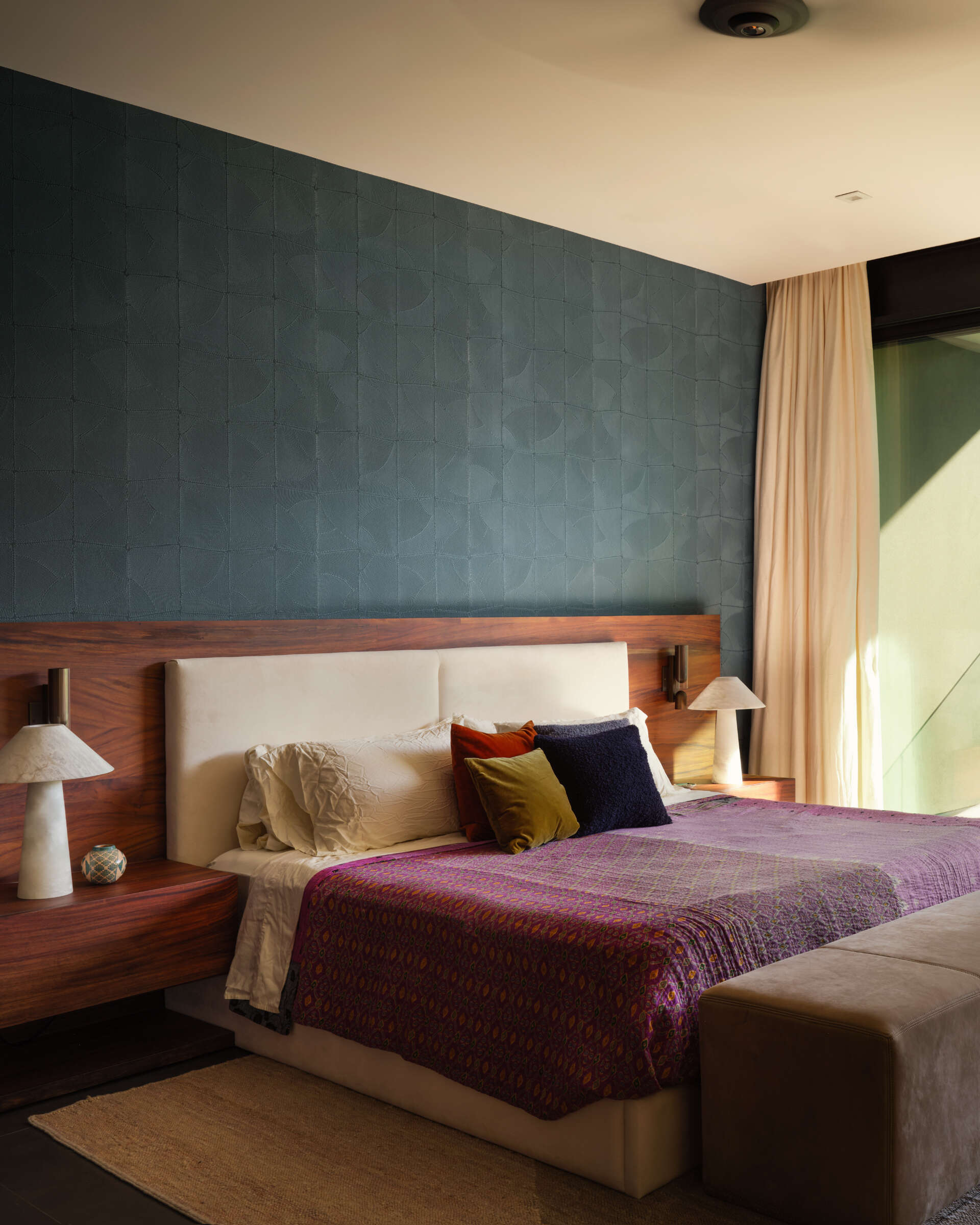 A textured blue feature wall brings depth to this bedroom, complemented by a wide timber headboard and softly upholstered bed. Plum bedding and jewel-toned cushions introduce color, while the restrained palette keeps the space feeling calm and inviting.