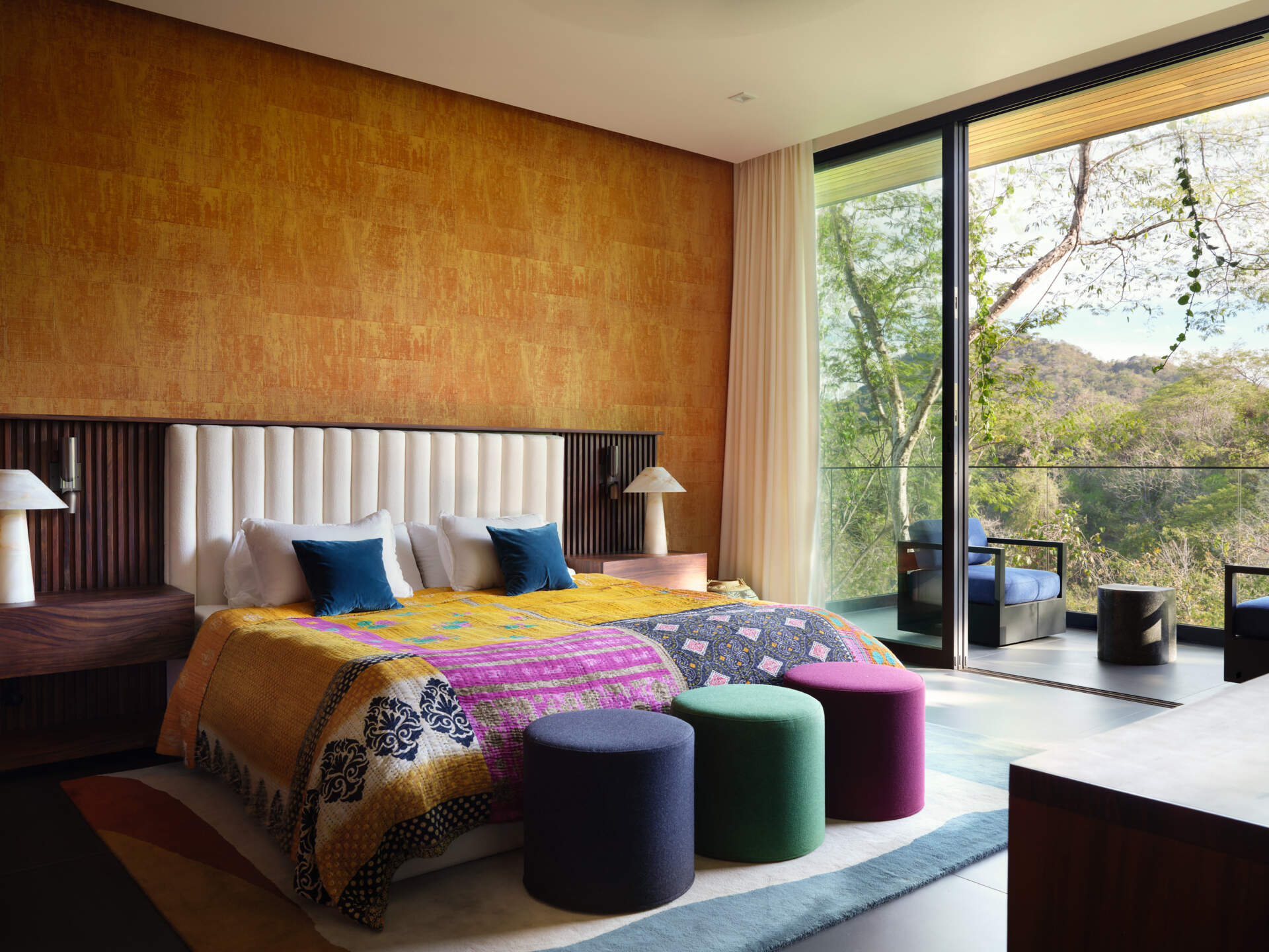 Earthy ochre walls pair with dark timber paneling in another bedroom. A colorful patchwork bedspread introduces layers of mustard, pink, and navy, echoed by the trio of upholstered stools positioned at the foot of the bed.