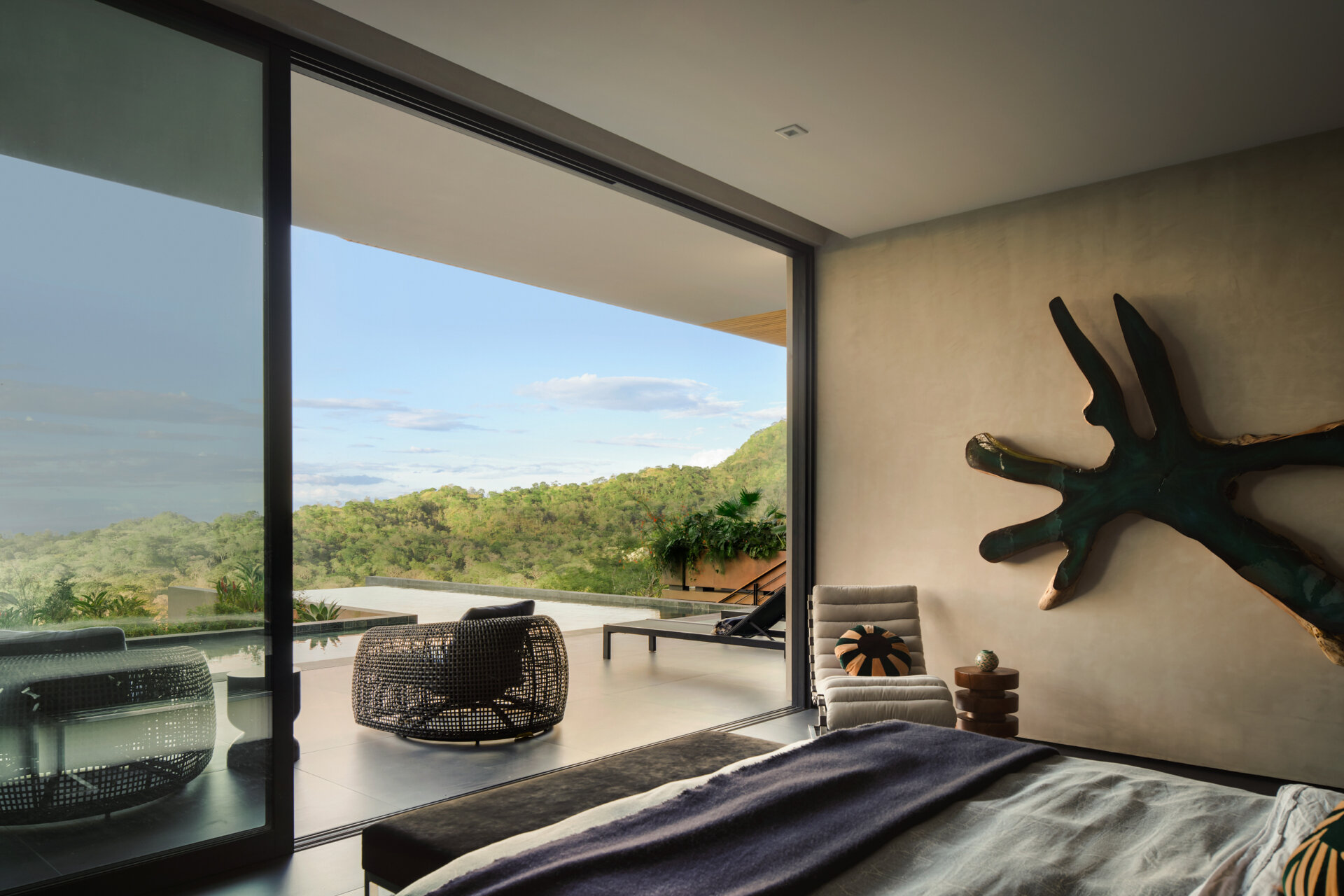 This bedroom opens directly onto a generous terrace, while a sculptural timber artwork provides a natural focal point against the plaster wall. Soft grey bedding, a low upholstered bench, and a comfortable lounge chair complete the room.