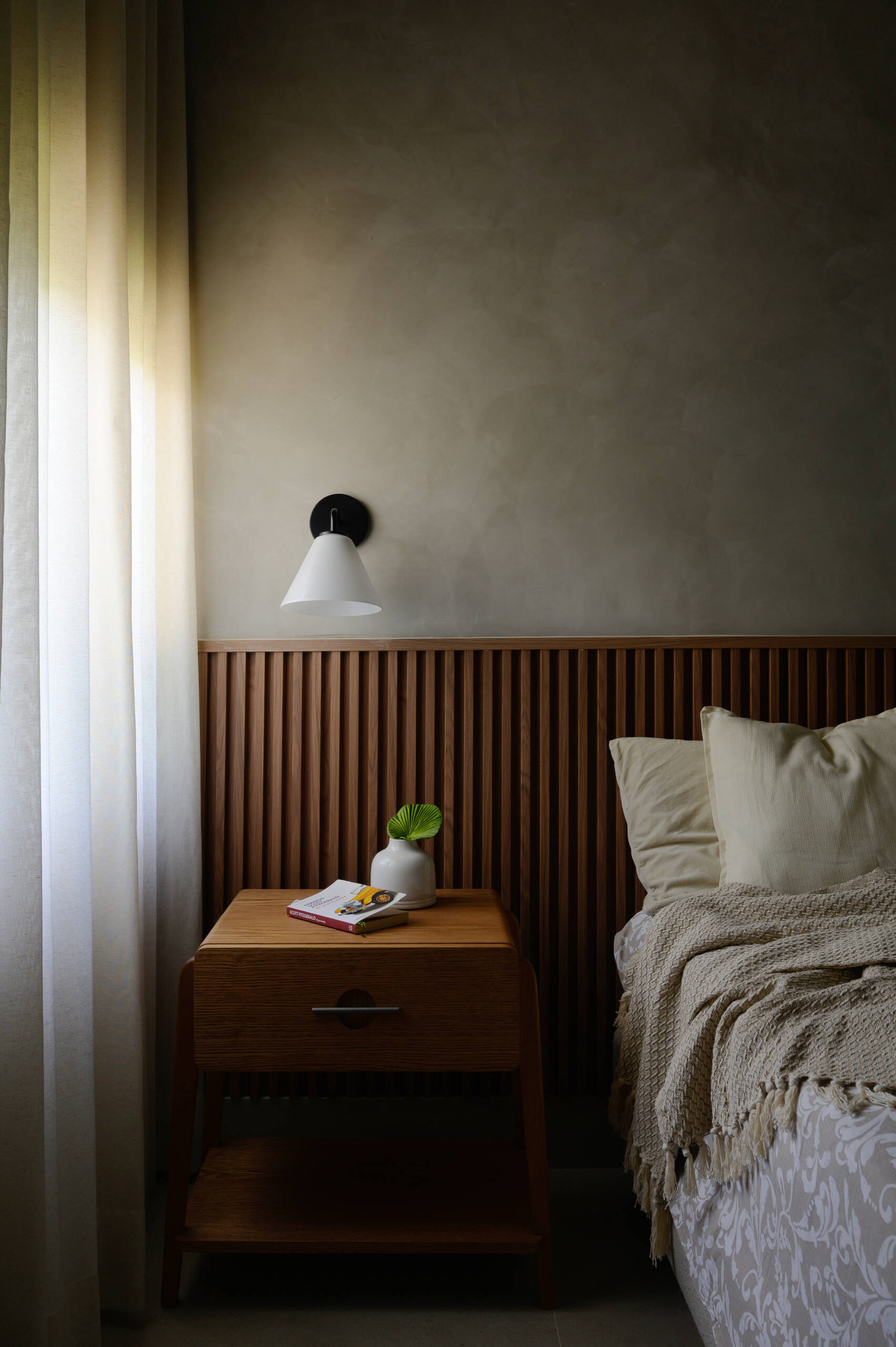 Discover a cosy modern bedroom with a full-width timber slat headboard, textured plaster walls, soft neutral furnishings and filtered natural light.