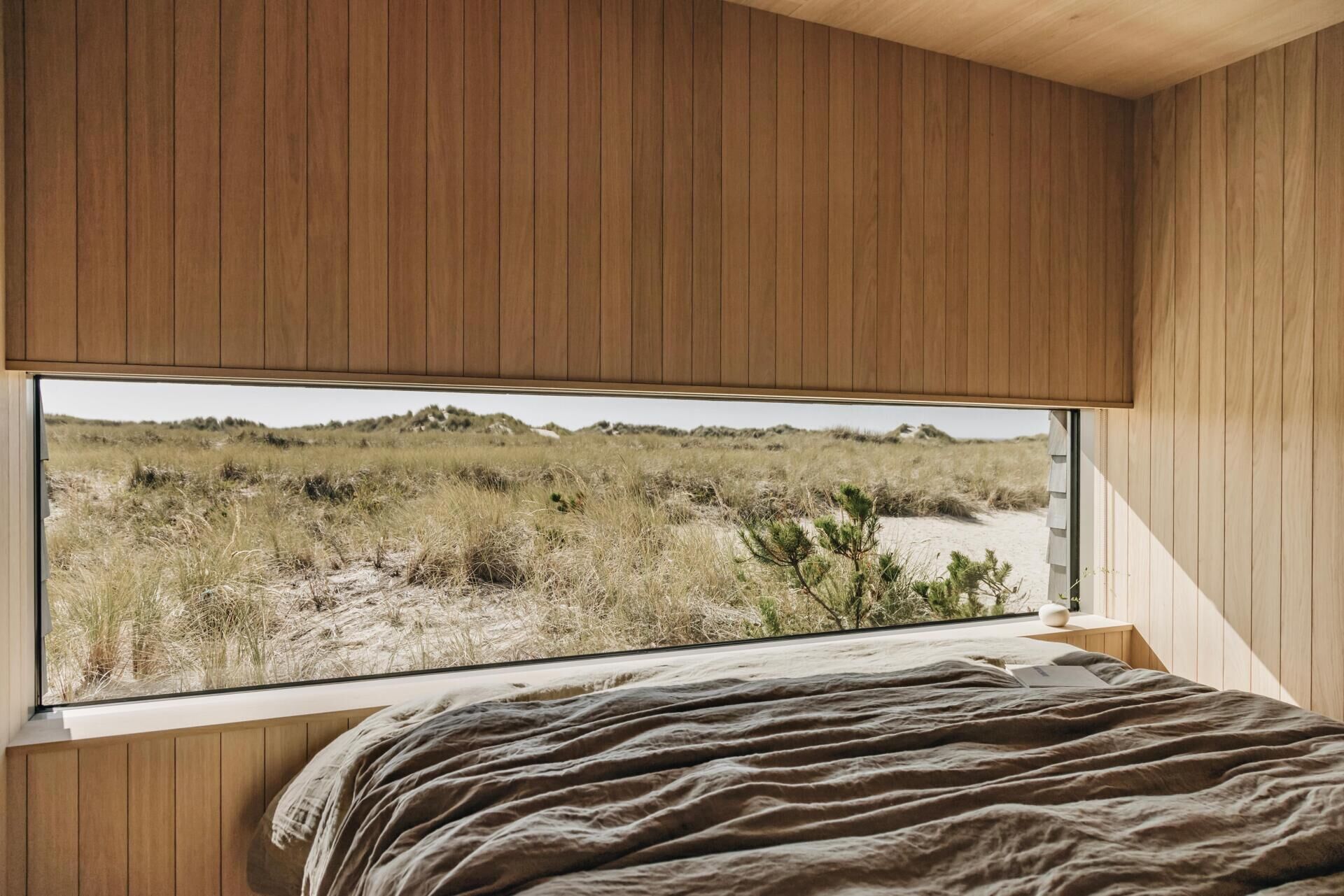 In the bedroom, a wide picture window sits just above the bed, making the changing grasses and sandy ridges the first thing seen each morning. The view feels uninterrupted, with slim window frames allowing the landscape to take centre stage.